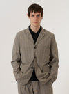 A model wears the Grey Brown Wales Glen Check Barra Jacket over a light-colored shirt, standing against a plain white background. The jacket features a subtle glen check pattern in grey and brown tones, three front buttons, and large patch pockets. The model is shown from the front, highlighting the garment's fit and structured collar.