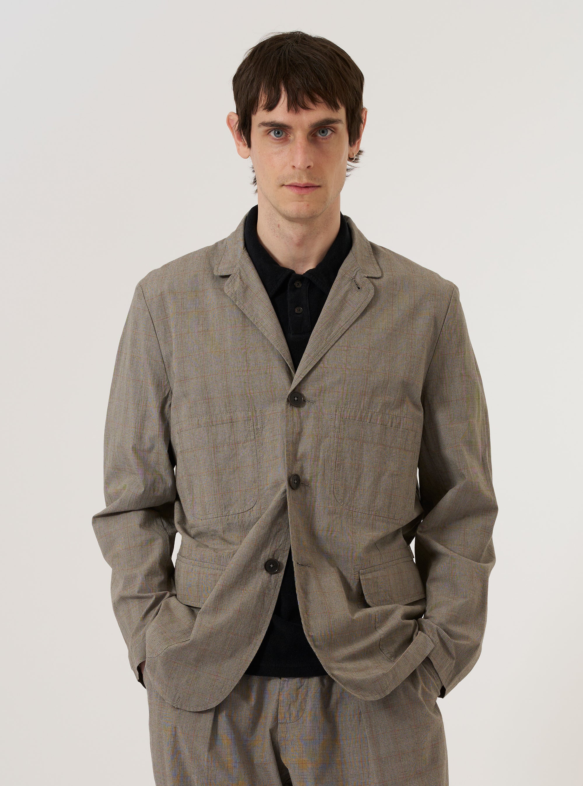 A model wears the Grey Brown Wales Glen Check Barra Jacket over a light-colored shirt, standing against a plain white background. The jacket features a subtle glen check pattern in grey and brown tones, three front buttons, and large patch pockets. The model is shown from the front, highlighting the garment's fit and structured collar.