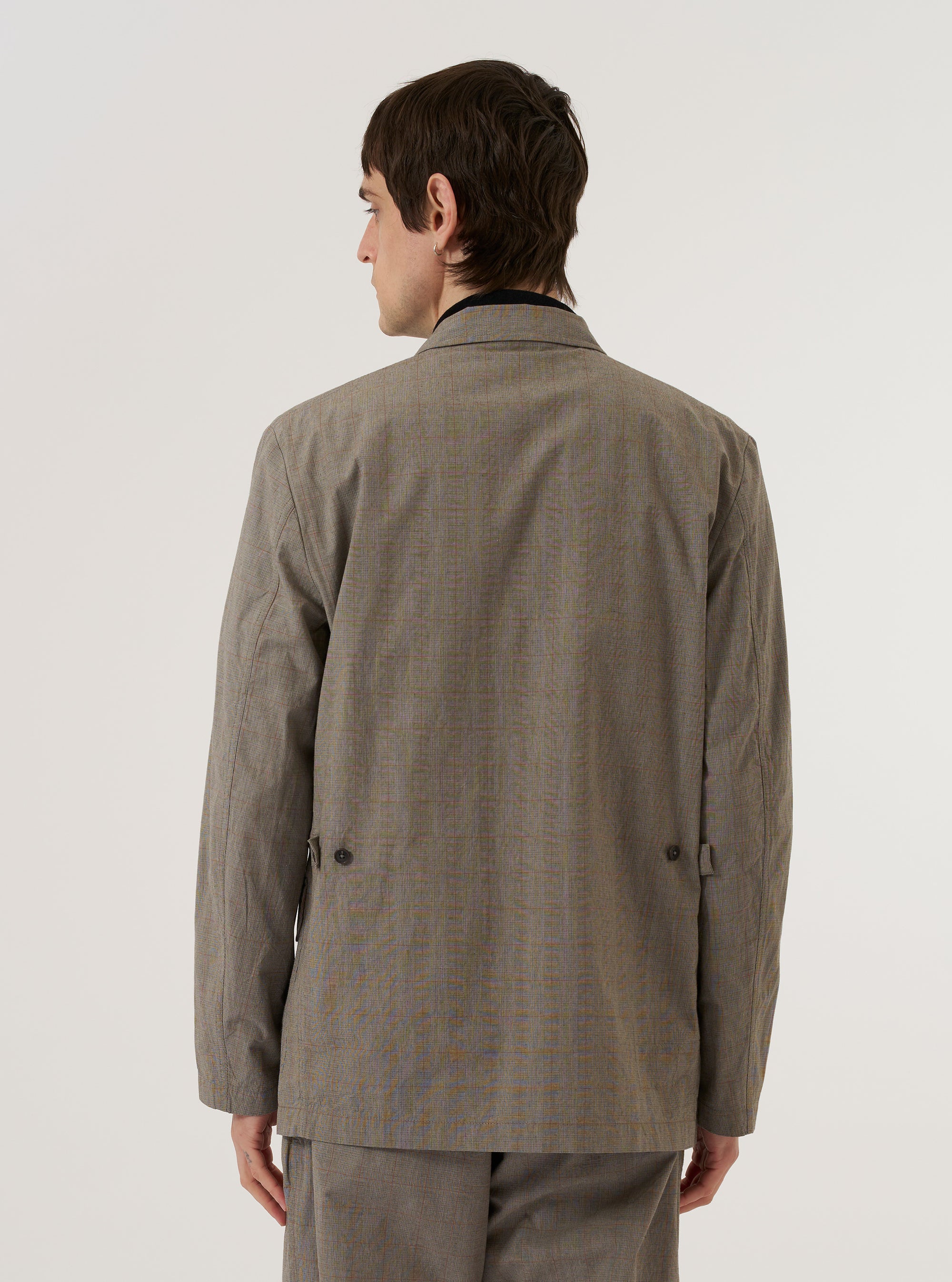 A model wears the Grey Brown Wales Glen Check Barra Jacket over a dark top against a plain white background. The image shows a rear view of the jacket, highlighting the center back vent and the drape of the glen check patterned fabric. The button waist adjusters on the sides are also visible.