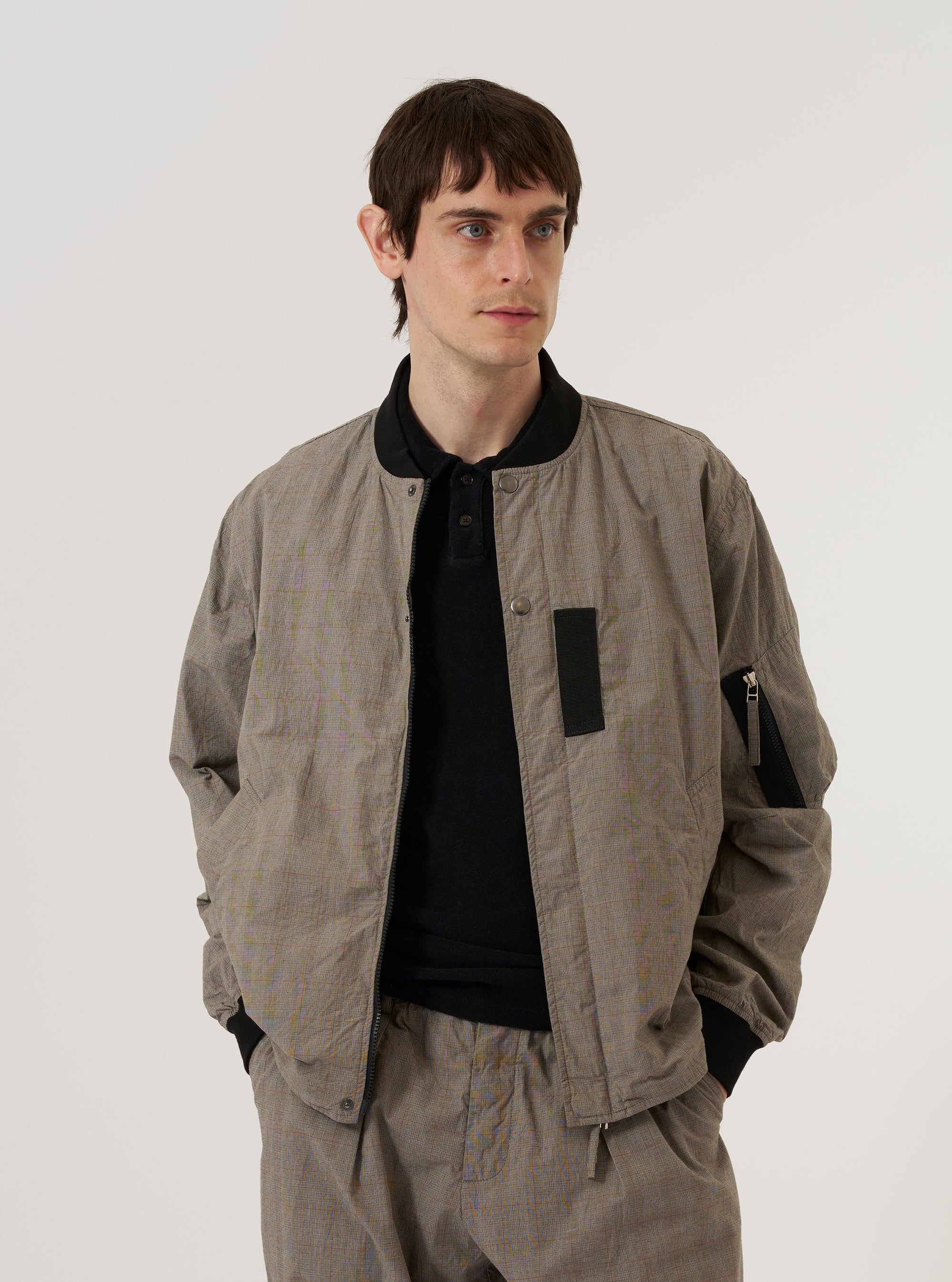 A model wears the Grey Brown Wales Glen Check NS Bomber Jacket over a dark top, standing against a plain white background. The jacket features a classic bomber silhouette with a central zip closure, a ribbed collar, and two slanted side pockets. The garment is characterized by its detailed grey and brown glen check plaid pattern throughout.