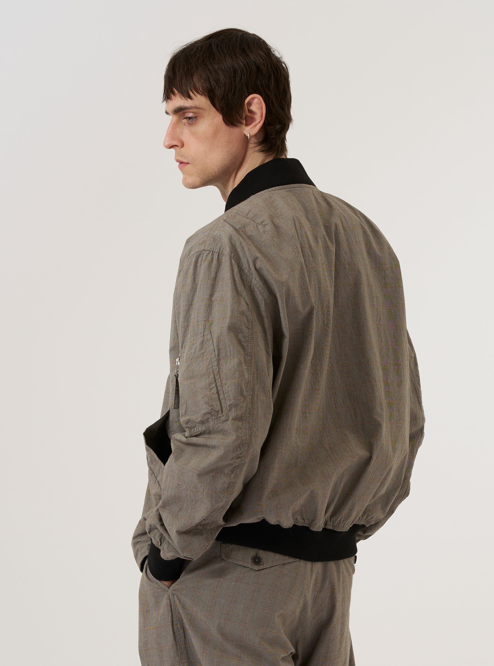 The Grey Brown Wales Glen Check NS Bomber Jacket is worn by a model against a plain white background. The image, captured from the rear, highlights the jacket's relaxed fit and the intricate grey and brown glen check pattern. The garment features a ribbed collar, cuffs, and hem, with a clean, straight-cut back panel. No mannequin is present in the image.
