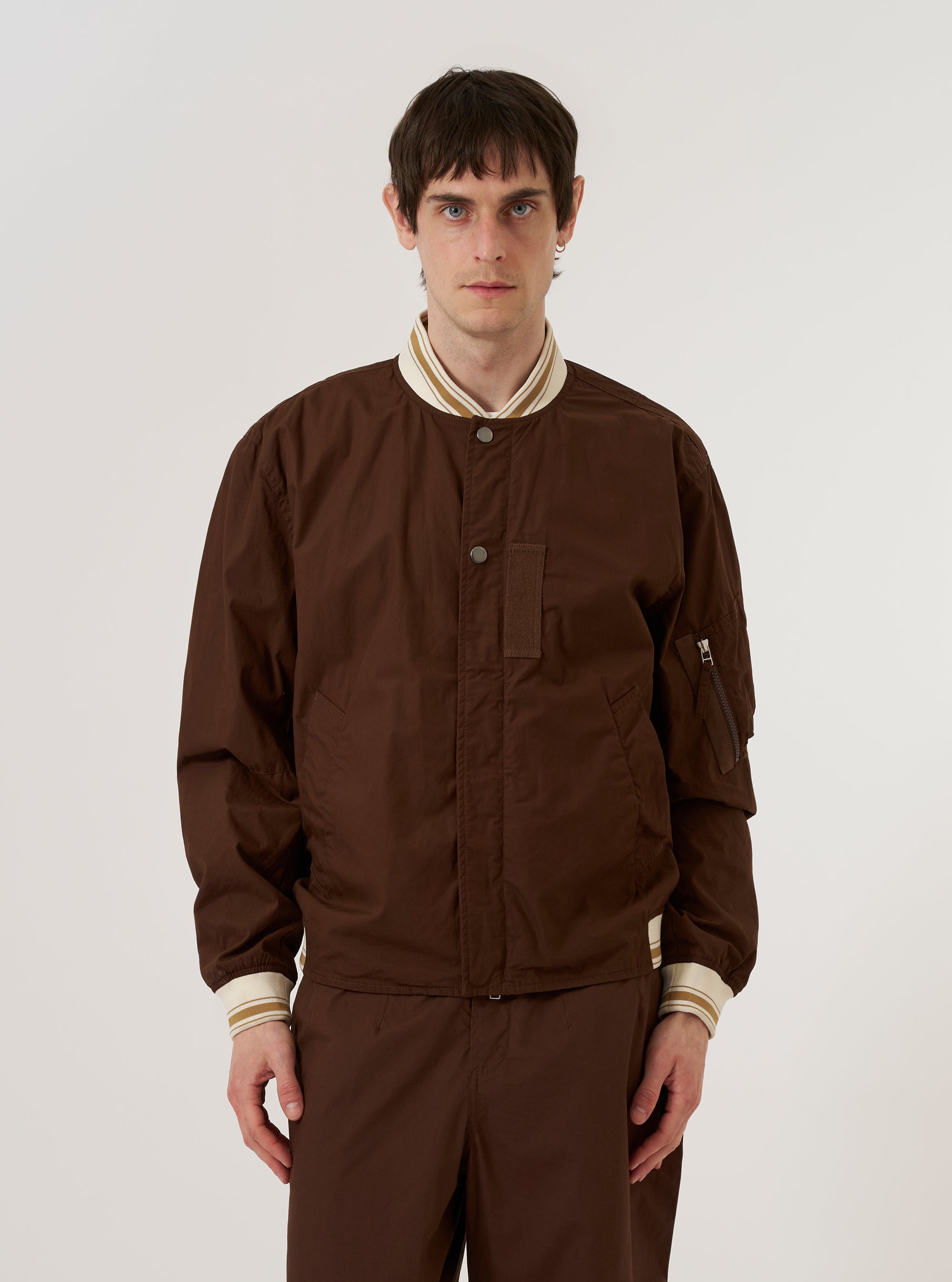 A model wears the Brown Peached Poplin NS Bomber Jacket against a plain, light grey background. The jacket features a front closure with both a zipper and metal pop studs, large hand warmer pockets, and contrast stripe ribbing at the collar and cuffs. The brown cotton poplin fabric has a smooth, matte finish. The model is shown from the chest up, highlighting the garment's regular fit and military-inspired design.