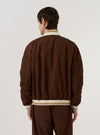 A model is wearing the Brown Peached Poplin NS Bomber Jacket over a light-colored top, shown from a rear angle against a plain white background. The jacket features articulated sleeves, a straight hem, and contrast stripe ribbing on the collar and cuffs. The brown cotton fabric has a smooth, peached finish.