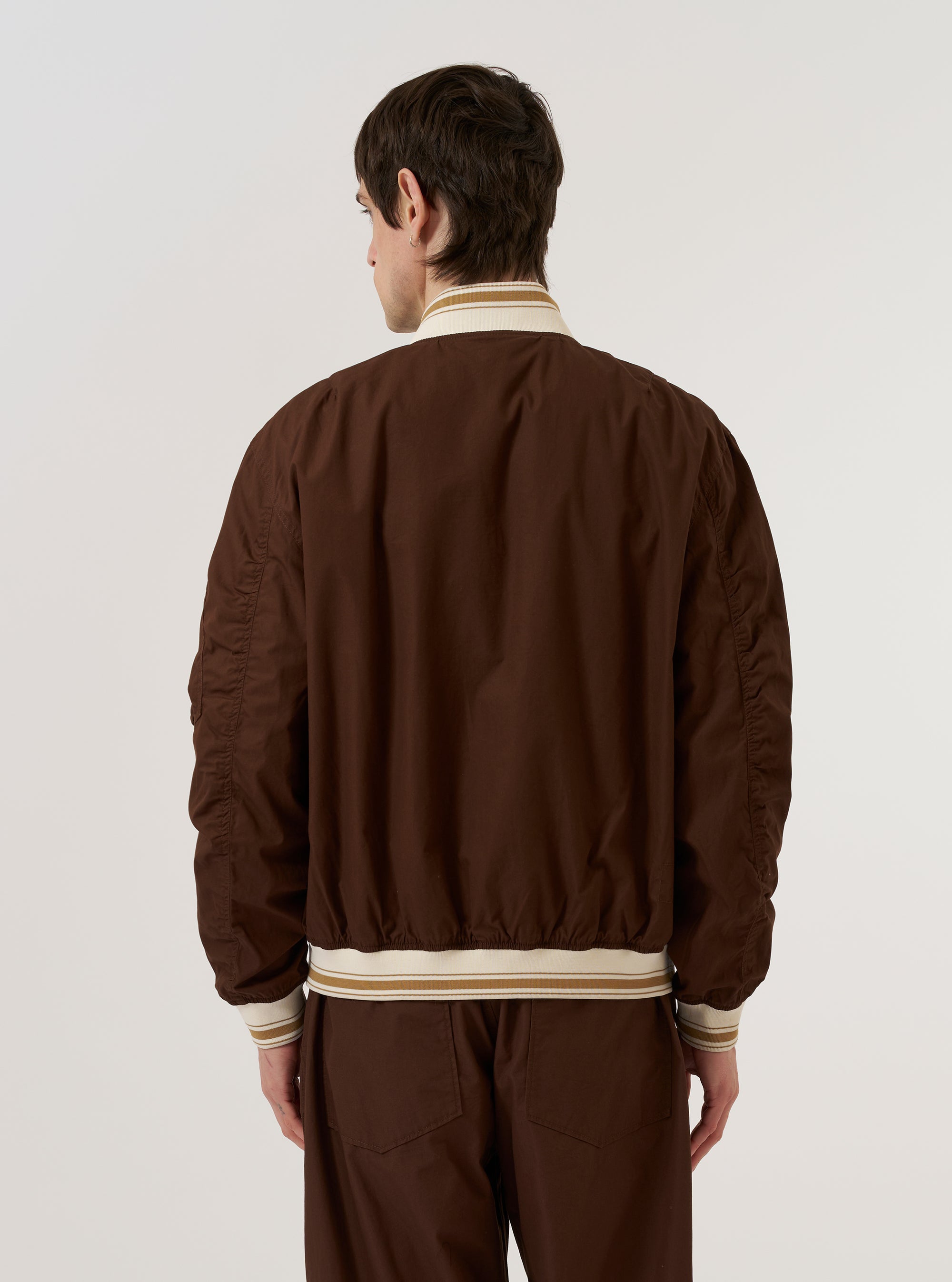 A model is wearing the Brown Peached Poplin NS Bomber Jacket over a light-colored top, shown from a rear angle against a plain white background. The jacket features articulated sleeves, a straight hem, and contrast stripe ribbing on the collar and cuffs. The brown cotton fabric has a smooth, peached finish.