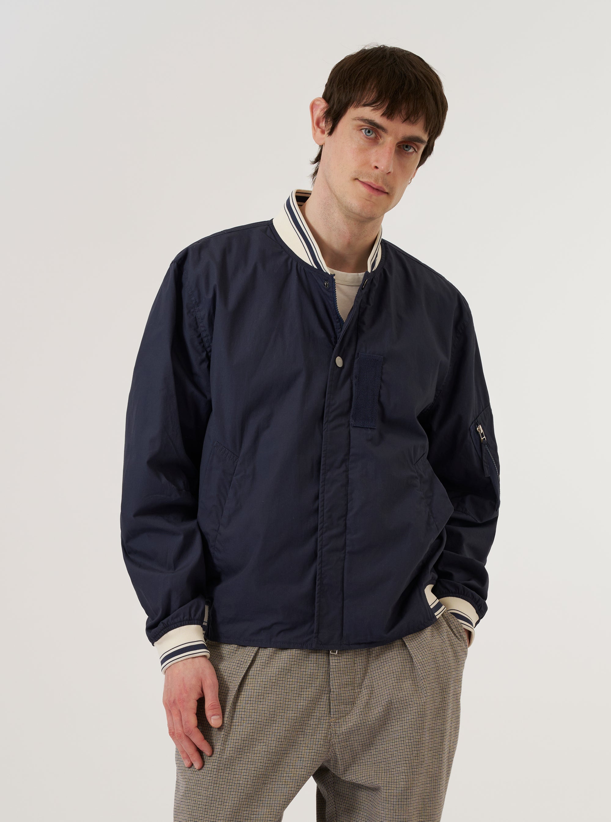 A model wears the Navy Peached Poplin NS Bomber Jacket over a light-colored shirt, standing against a plain, neutral background. The jacket features a partially concealed two-way zip front closure with metal pop studs, large hand warmer pockets, and contrast stripe ribbing at the collar, cuffs, and hem. The navy peached poplin fabric has a smooth, matte finish.