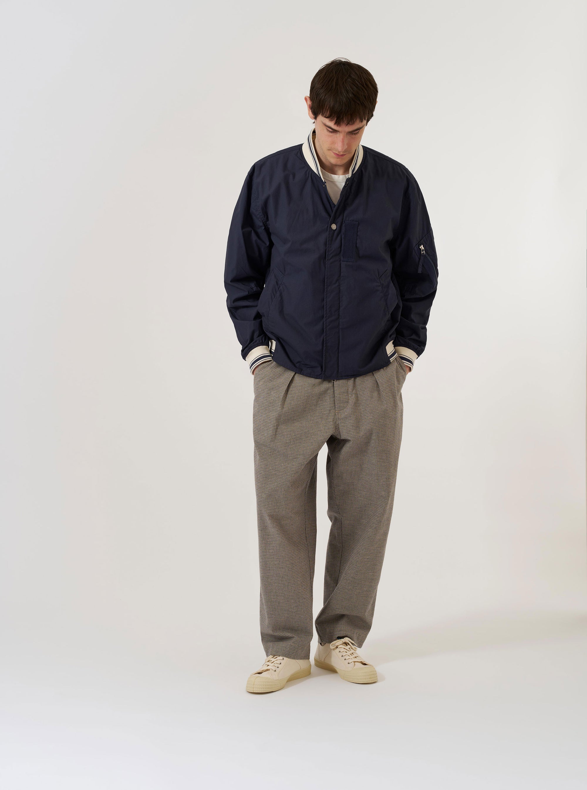 The Navy Peached Poplin NS Bomber Jacket is shown as a flat lay against a white background. The jacket features a central two-way zip front closure with visible metal pop studs, contrast stripe ribbing on the collar and cuffs, and large hand warmer pockets. A zipped utility stash pocket is visible on the sleeve, and the garment is constructed with articulated sleeves.