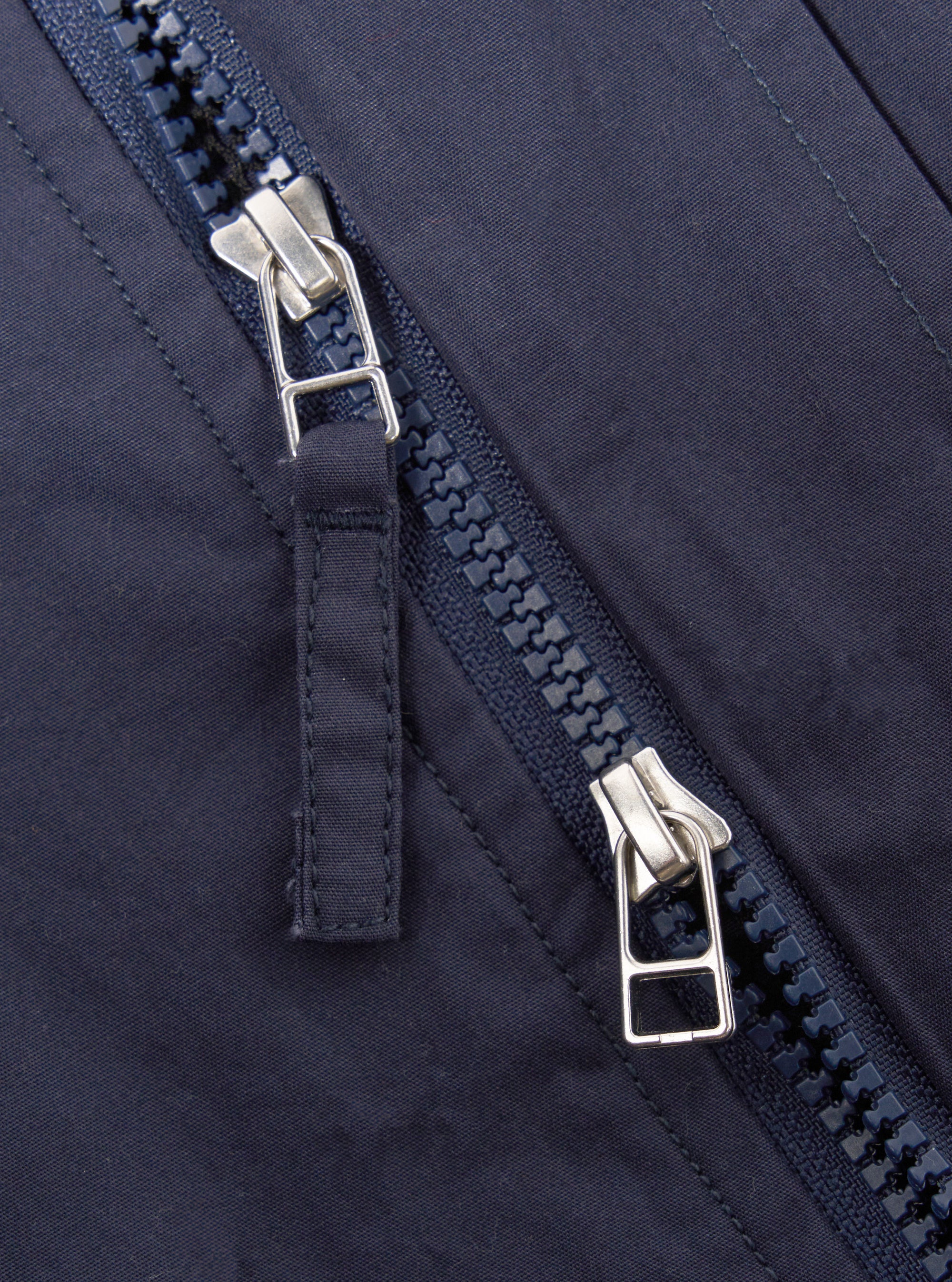 The Navy Peached Poplin NS Bomber Jacket is shown as a flat lay against a plain white background. The jacket features a central zip front with a buttoned placket, a ribbed collar, and a utility zip pocket on the left sleeve. Two large hand warmer pockets are visible on the front, and the fabric has a smooth, matte finish.