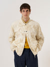 A model wears the Ecru 3D Flower NS Bomber Jacket over a light-colored shirt, standing against a plain white background. The jacket features a distinct 3D floral texture throughout the ecru fabric, contrast ribbed trim at the collar and cuffs, and a visible zippered utility pocket on the sleeve. The front closure includes a two-way zip and metal pop studs.
