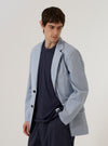 A model wears the Navy Kennedy Stripe Boston Blazer over a light-colored shirt, standing against a plain, neutral background. The blazer features notched lapels, a three-button front closure, and three patch pockets on the front. The fabric is a navy blue cotton with a fine white pinstripe pattern throughout.