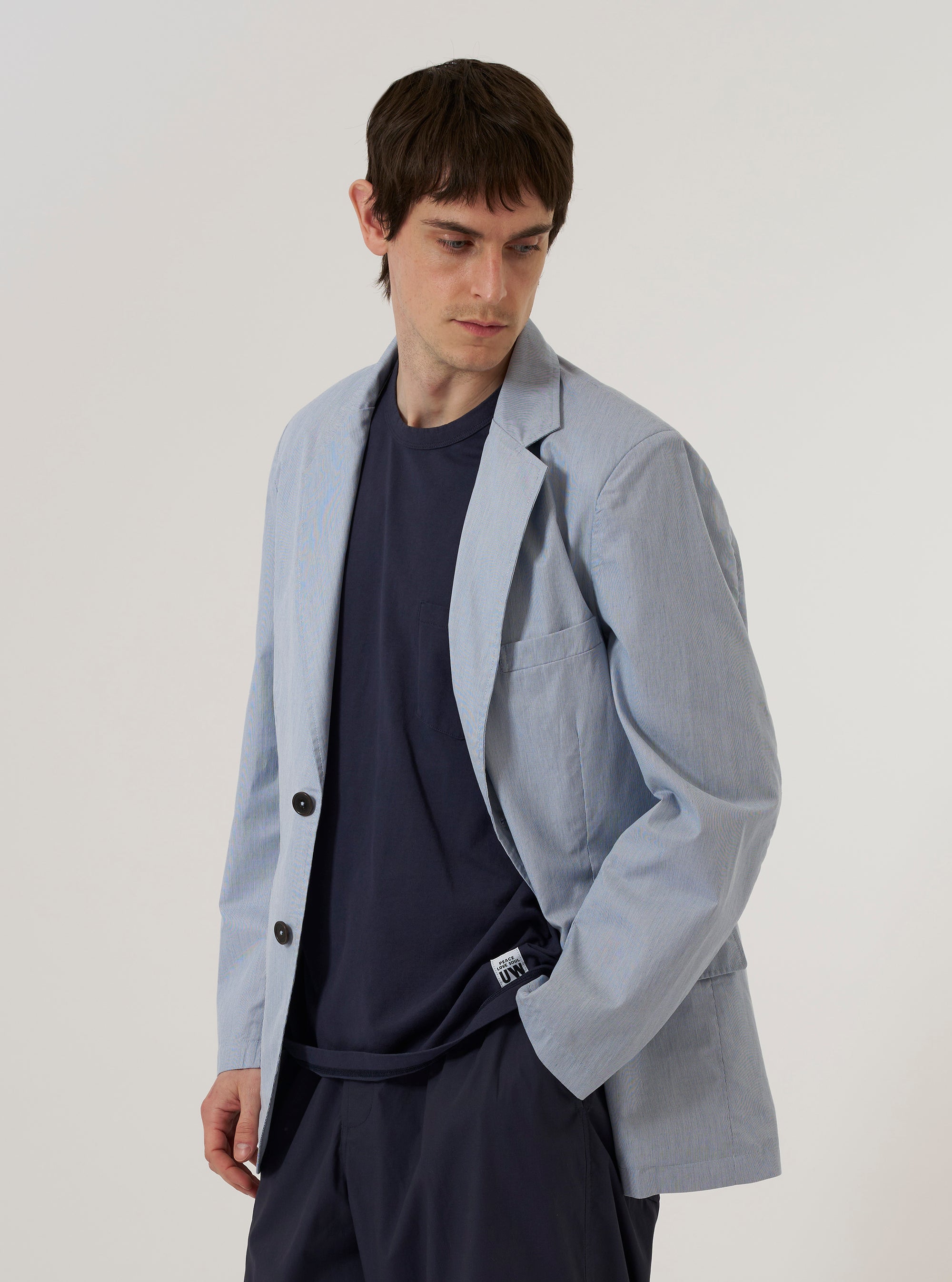 A model wears the Navy Kennedy Stripe Boston Blazer over a light-colored shirt, standing against a plain, neutral background. The blazer features notched lapels, a three-button front closure, and three patch pockets on the front. The fabric is a navy blue cotton with a fine white pinstripe pattern throughout.