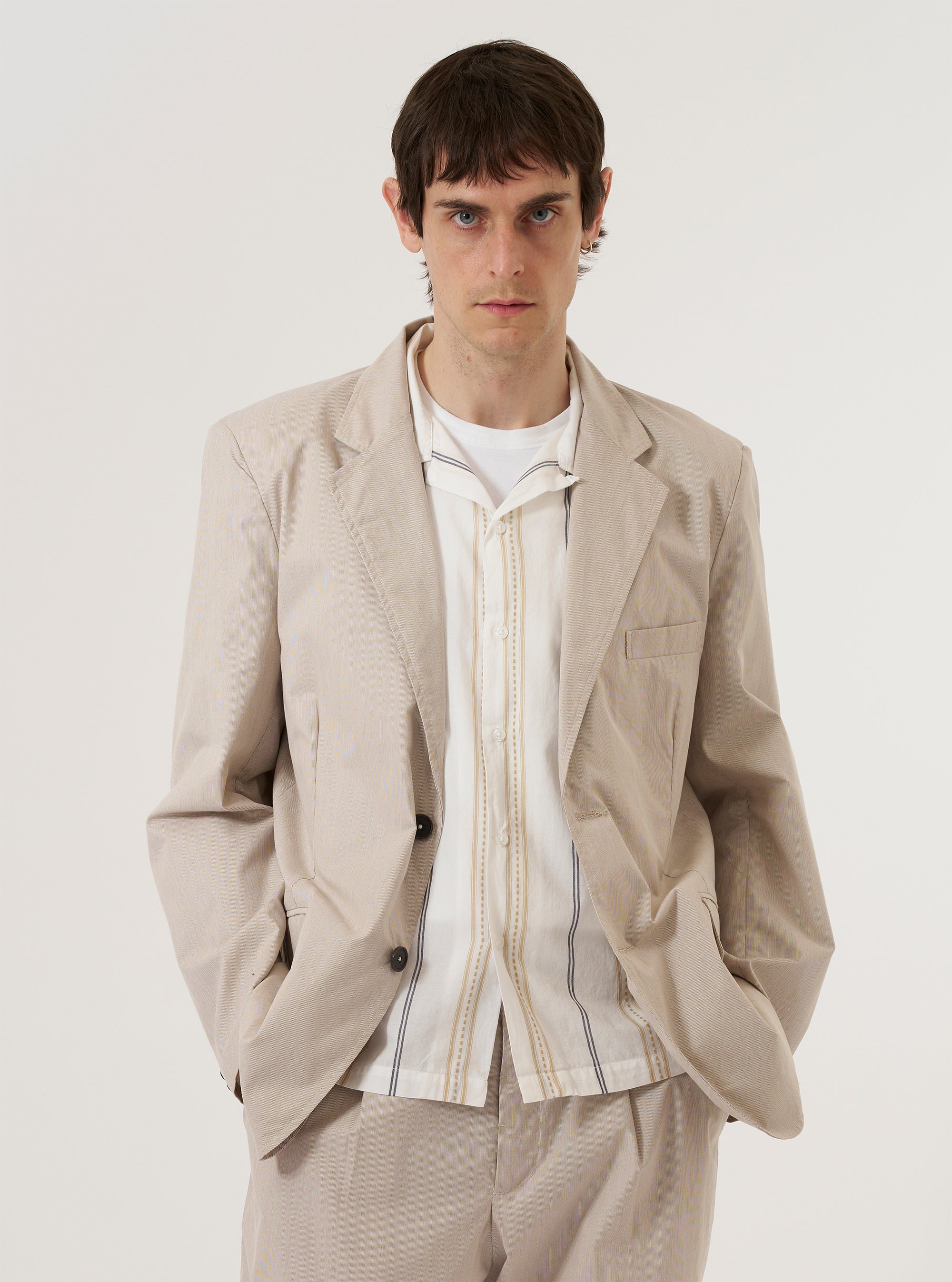 A model wears the Brown Kennedy Stripe Boston Blazer over a dark top, standing against a plain, light-colored background. The blazer features a notched lapel, a three-button front closure, and two large patch pockets at the waist. The brown fabric is characterized by a distinct vertical stripe pattern throughout.