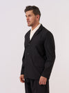 A model wears the Black Recycled Polytech Capitol Jacket against a plain white background. The jacket features a three-button front closure, a notched lapel, and three front patch pockets including one at the chest and two at the waist. The garment is shown from a front angle, highlighting its relaxed fit and the smooth, technical finish of the black recycled fabric. The model layers the jacket over a light-colored top and dark trousers.