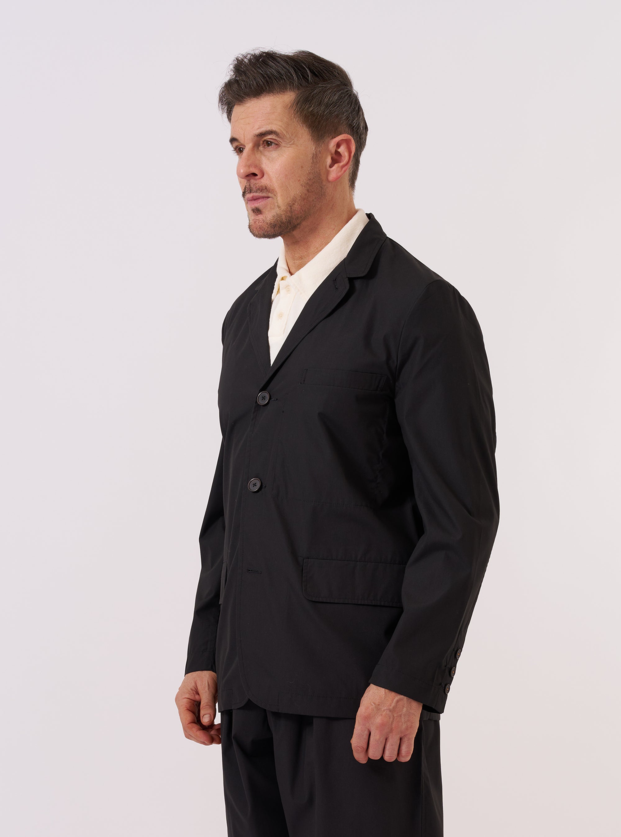 A model wears the Black Recycled Polytech Capitol Jacket against a plain white background. The jacket features a three-button front closure, a notched lapel, and three front patch pockets including one at the chest and two at the waist. The garment is shown from a front angle, highlighting its relaxed fit and the smooth, technical finish of the black recycled fabric. The model layers the jacket over a light-colored top and dark trousers.