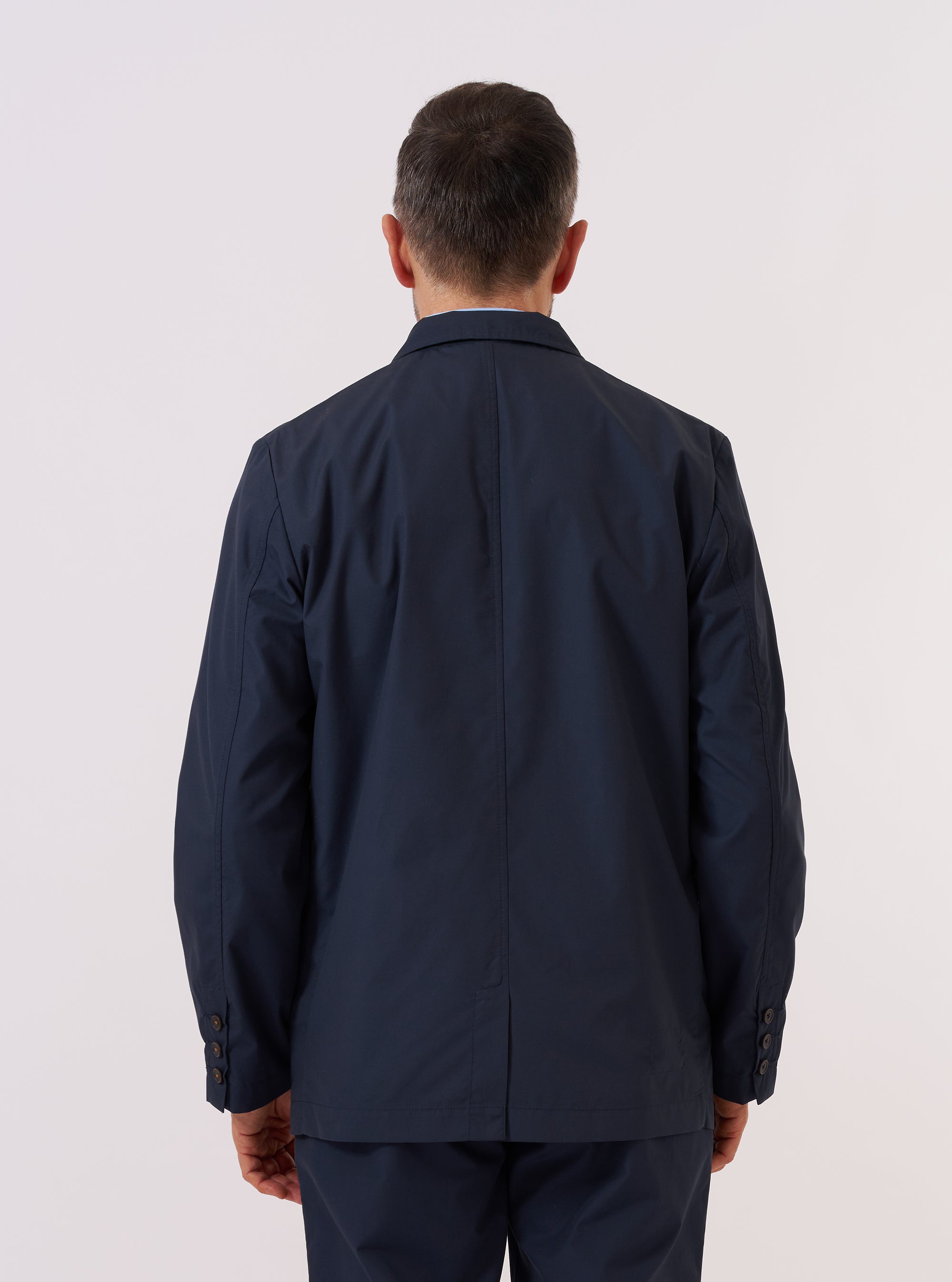 A model wears the Navy Recycled Polytech Capitol Jacket over a light-colored top, shown from the back against a plain white background. The jacket features a clean, structured silhouette with a central seam and a small brand tag at the side. The model is standing with their arms at their sides.