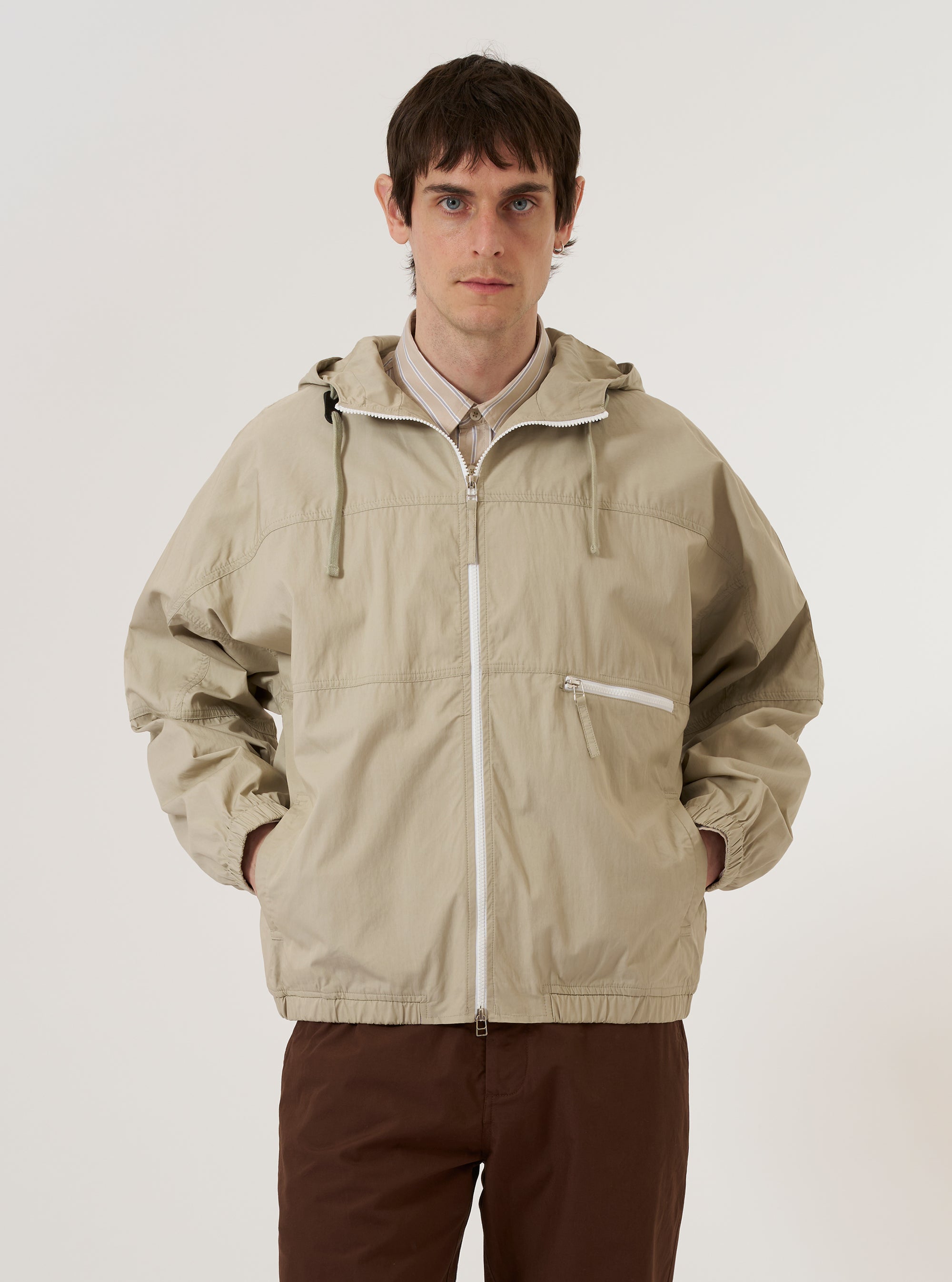 A model wears the Sand Cotton Recycled Nylon Mix Daley Jacket against a plain white background. The jacket is a light sand color and features a large hood, a full-length front zipper, and two oversized patch pockets at the waist. The model is shown from a front angle, highlighting the jacket's relaxed fit and technical fabric texture.