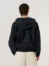 A model wears the Navy Cotton Recycled Nylon Mix Daley Jacket, shown from a rear three-quarter angle against a plain, light grey background. The image highlights the jacket's hooded design, a straight hem, and the smooth texture of the navy blue cotton-nylon blend fabric. The model is wearing the jacket over a light-colored top and dark trousers.
