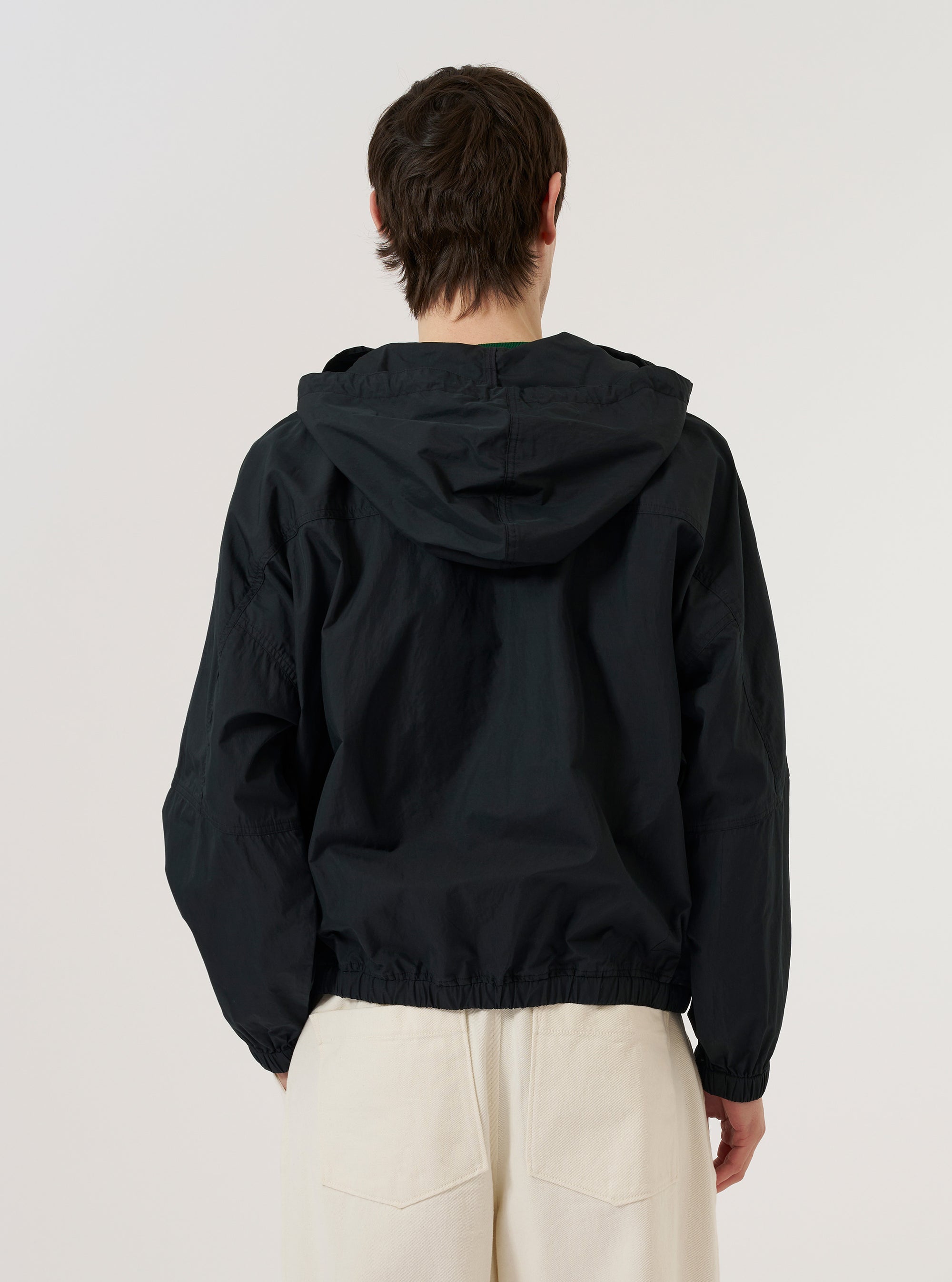 A model wears the Navy Cotton Recycled Nylon Mix Daley Jacket, shown from a rear three-quarter angle against a plain, light grey background. The image highlights the jacket's hooded design, a straight hem, and the smooth texture of the navy blue cotton-nylon blend fabric. The model is wearing the jacket over a light-colored top and dark trousers.