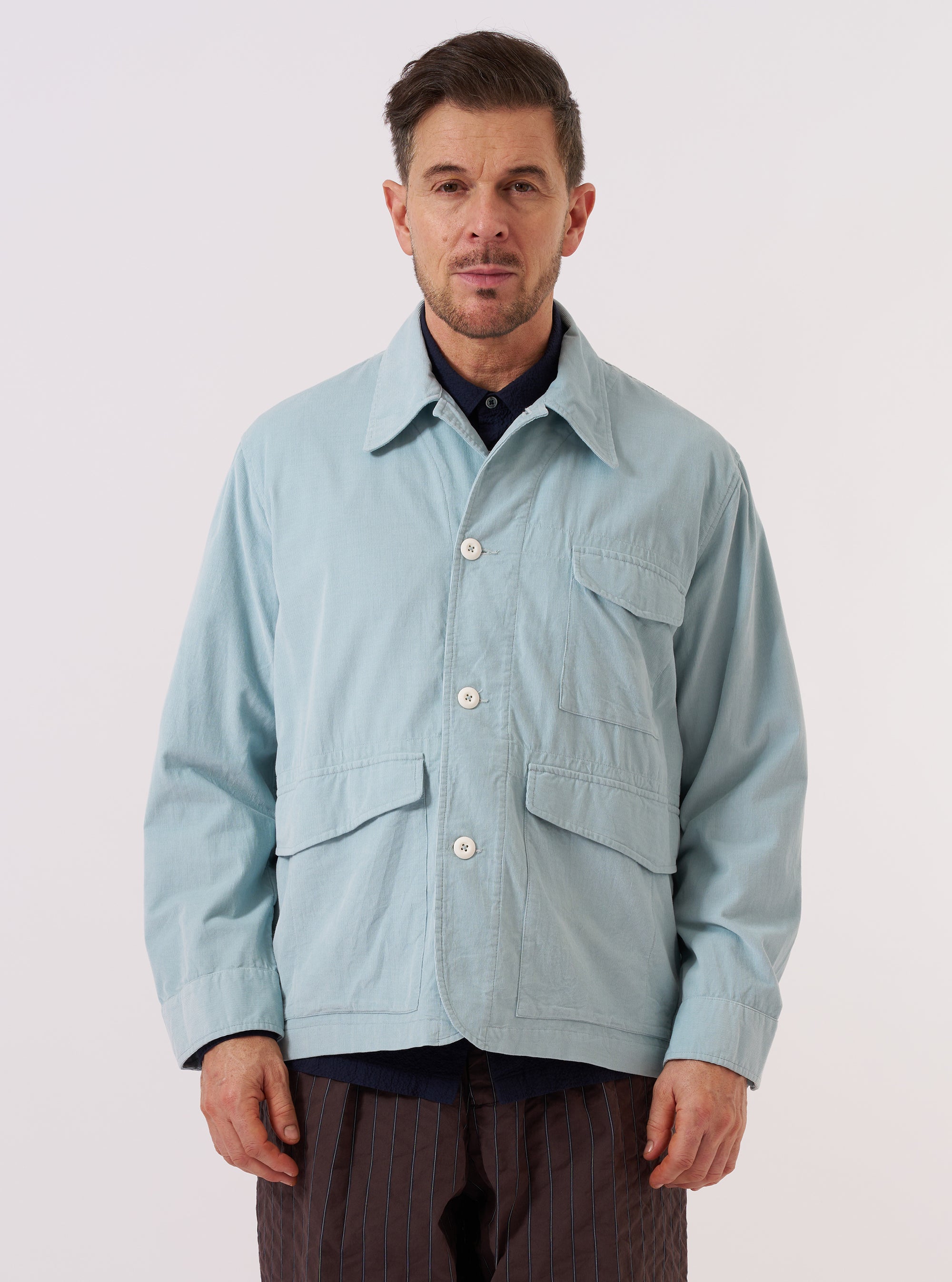 A model wears the Aqua Organic Fine Cord Duty Jacket over a white t-shirt, standing against a plain white background. The light blue corduroy jacket is shown from the front and features a classic collar, a button-down front, and three visible patch pockets on the torso. The model's hands are at their sides.