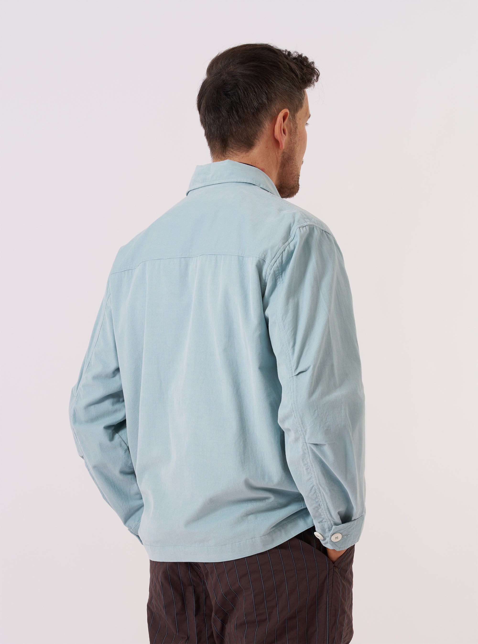 The Aqua Organic Fine Cord Duty Jacket is shown from the back, worn by a model against a plain white background. The image highlights the straight hem, a single vertical seam down the center of the back, and the fine-ribbed texture of the aqua-colored corduroy fabric. The model's hands are visible at their sides.