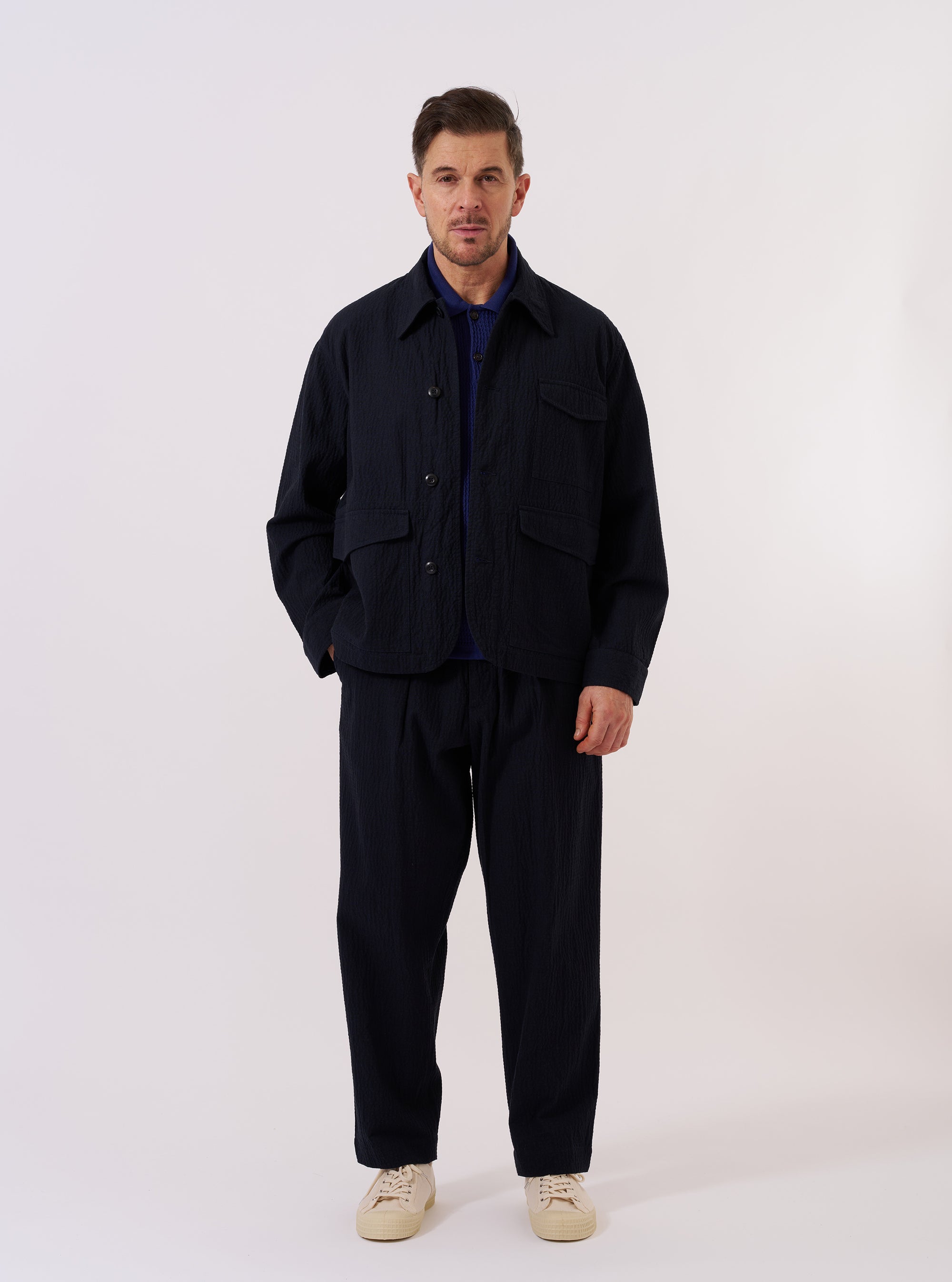 The Dark Navy Ospina Cotton Duty Jacket is shown as a flat lay against a white background. The jacket features two double-flap hip pockets, a single chest patch pocket, and a central button-down closure. The dark navy fabric has a visible seersucker texture, and the garment is designed with a curved hem and a structured collar.