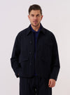 A model wears the Dark Navy Ospina Cotton Duty Jacket over a light-colored top, standing against a plain, neutral background. The jacket features a button-front closure, a structured collar, and multiple large patch pockets on the front. The fabric displays a subtle, puckered seersucker texture characteristic of the Ospina cotton.