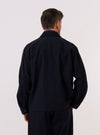 A model wears the Dark Navy Ospina Cotton Duty Jacket over a light-colored shirt, shown from a rear three-quarter angle against a plain, neutral studio background. The jacket features a structured collar and a straight hem, highlighting the distinct puckered texture of the dark navy seersucker fabric.