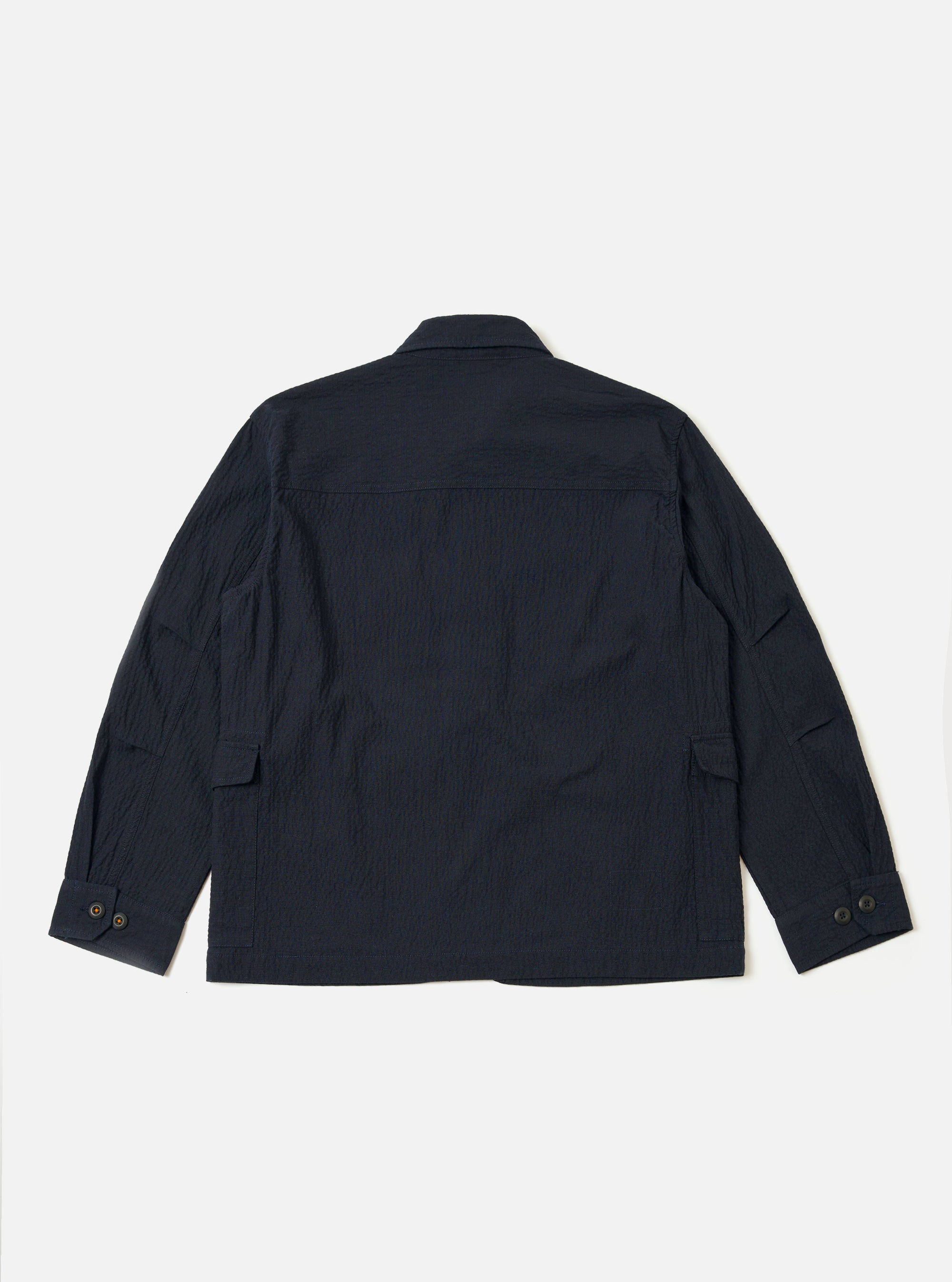 The Dark Navy Ospina Cotton Duty Jacket is shown from the back as a flat lay against a plain white background. The image highlights the jacket's curved hem, loose sleeves, and the puckered seersucker texture of the dark navy fabric.