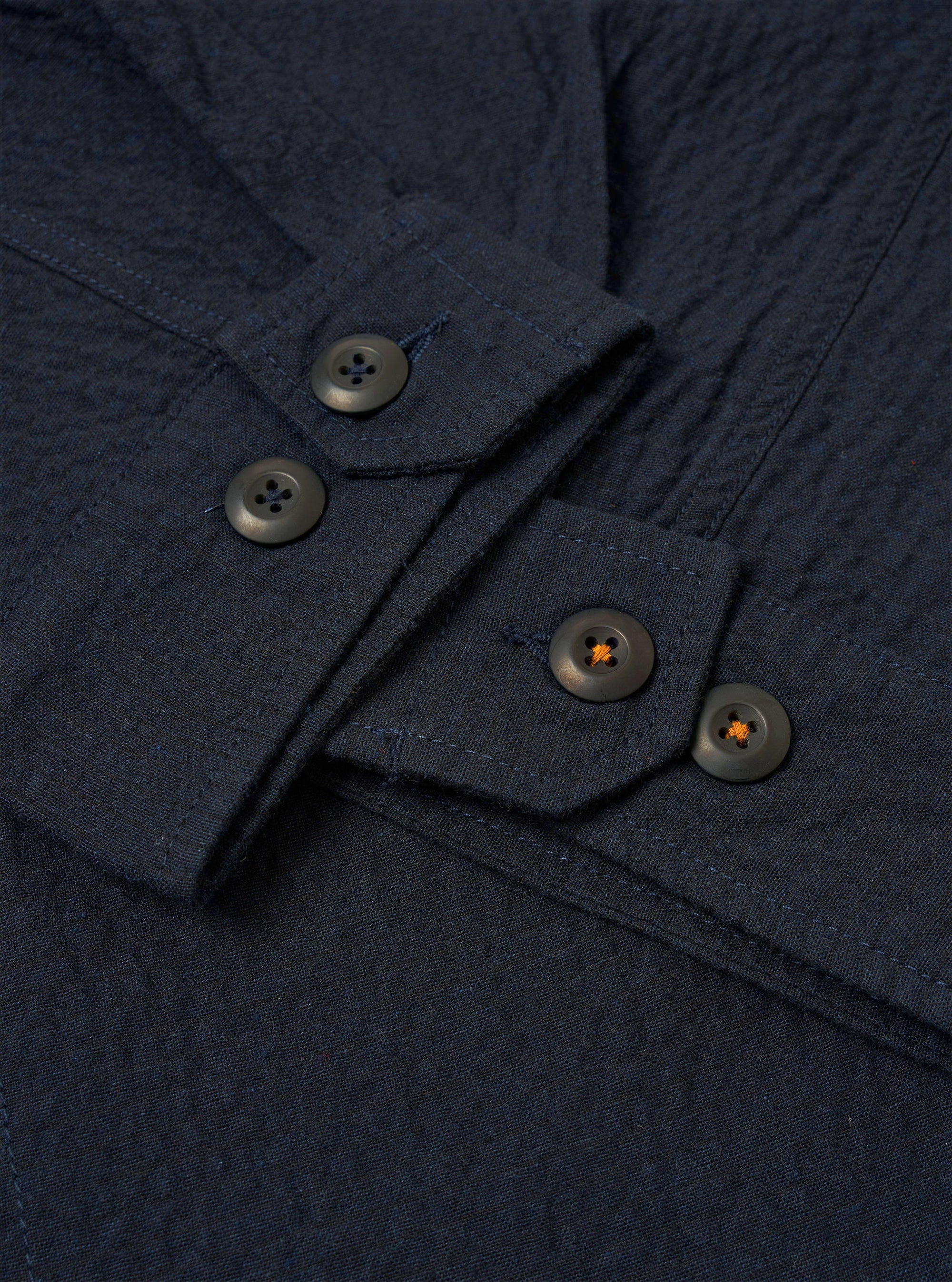 The Dark Navy Ospina Cotton Duty Jacket is shown from the back as a flat lay against a plain white background. The image highlights the clean construction of the shoulders and the straight hem of the jacket. The dark navy fabric features a subtle, puckered seersucker texture throughout.