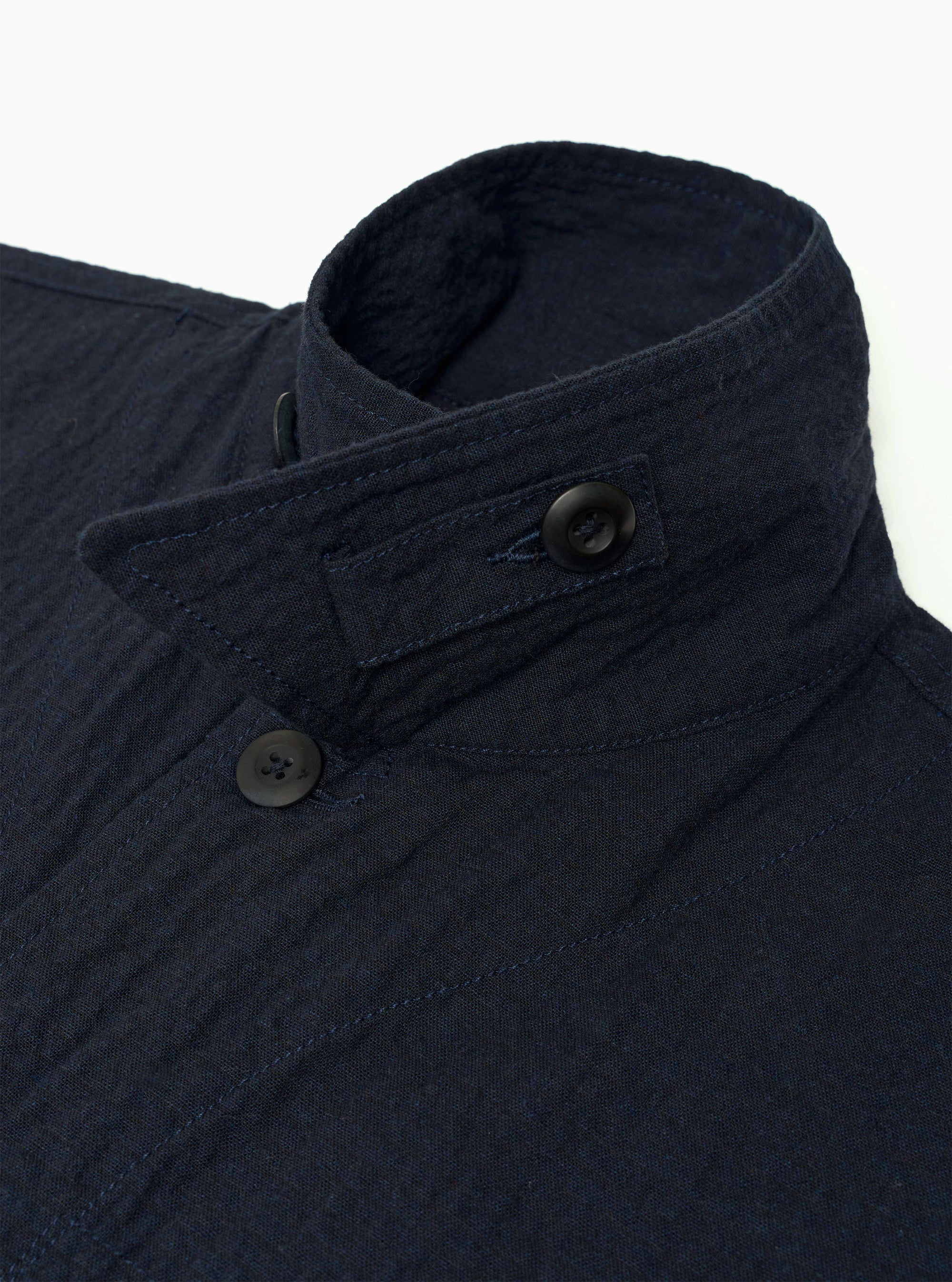 The Dark Navy Ospina Cotton Duty Jacket is shown from a rear three-quarter angle as a flat lay against a plain white background. The image highlights the jacket's oversized fit, loose darted sleeves, and curved hem. The dark navy fabric features a visible puckered seersucker texture.