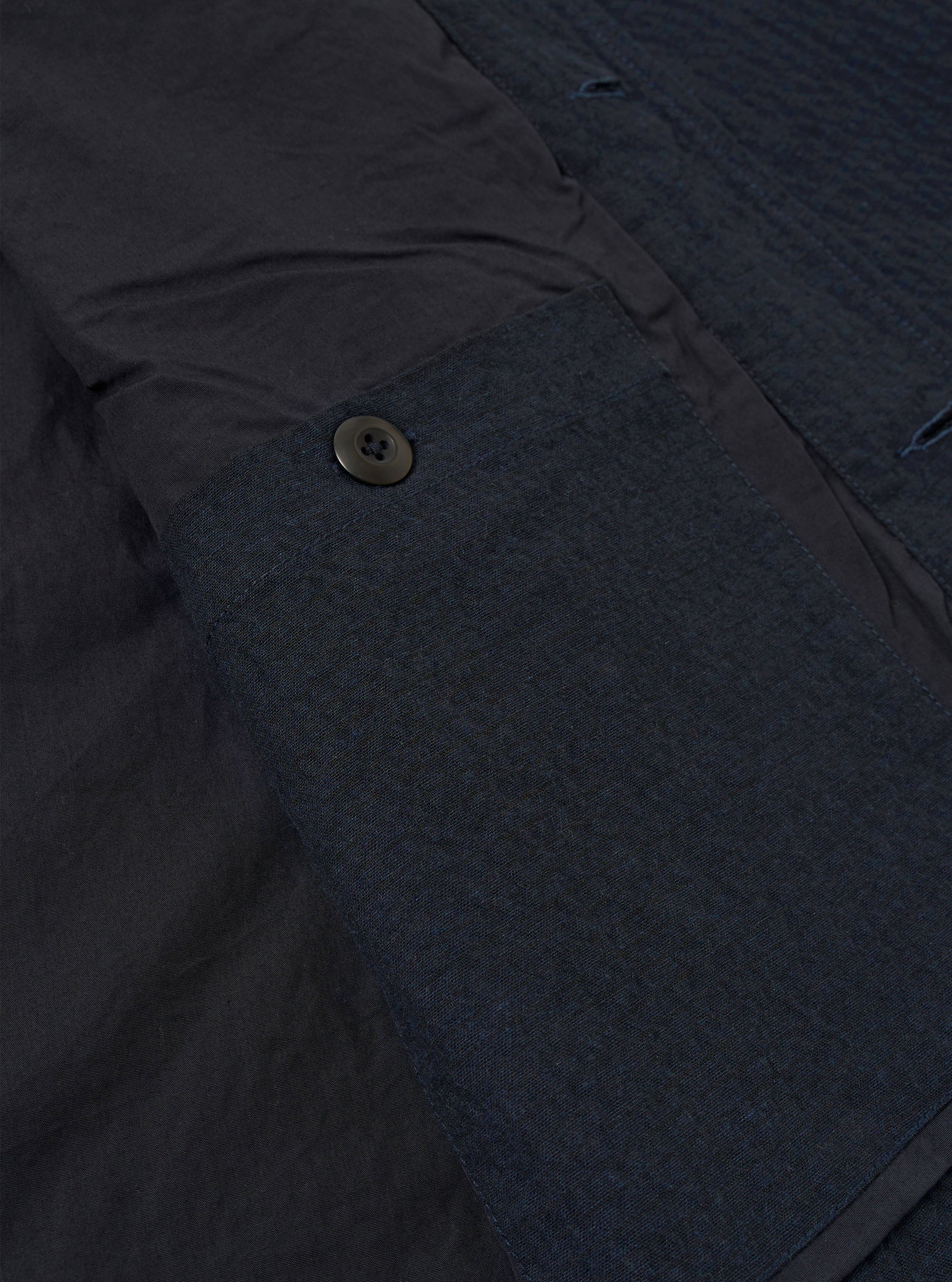 The Dark Navy Ospina Cotton Duty Jacket is shown in a detail view as a flat lay against a plain white background. The image highlights the textured seersucker finish of the dark navy fabric, the clean stitching of the hem, and the curved shape of the lower garment.