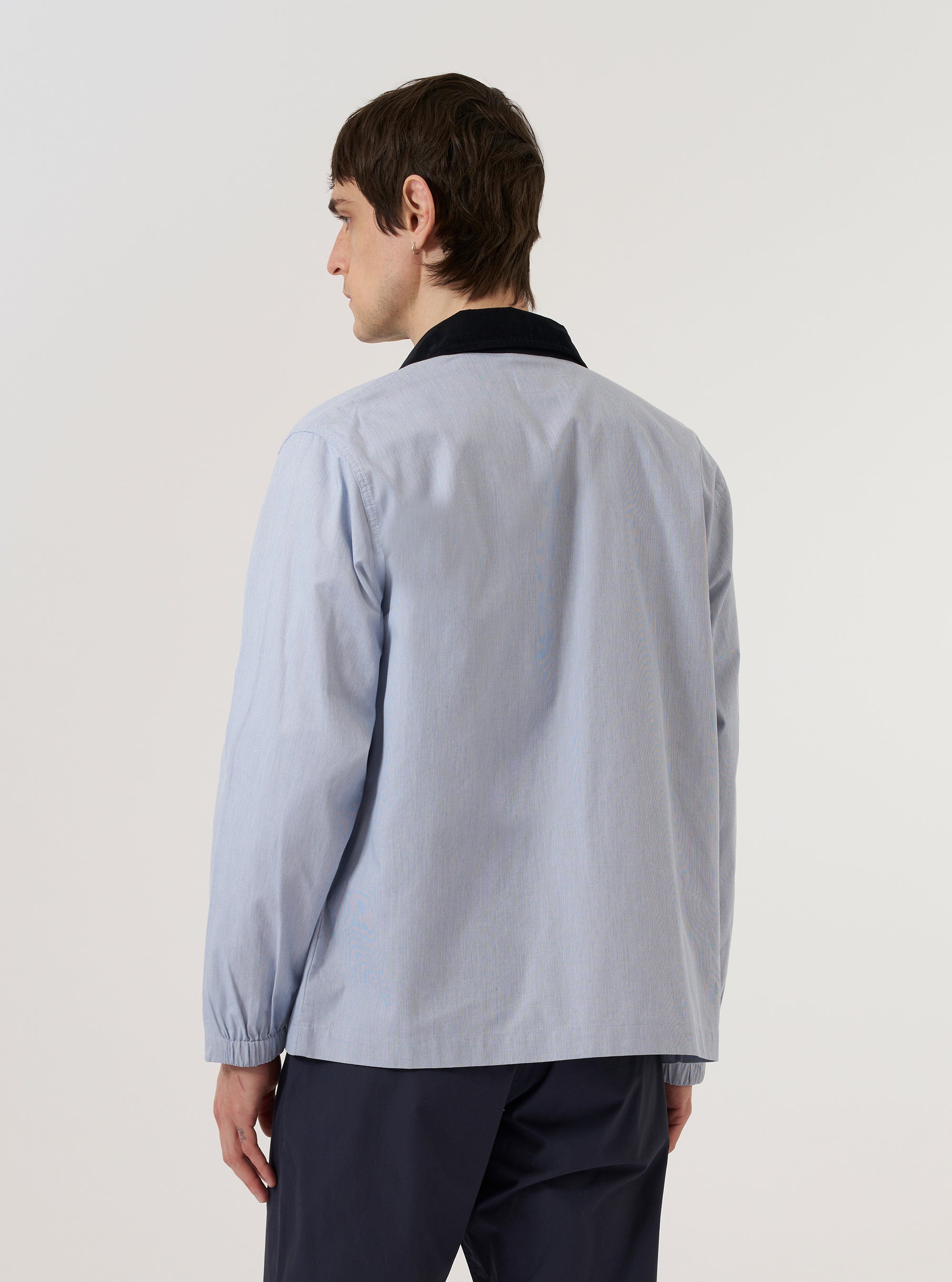 A model wears the Navy Kennedy Stripe Labour Jacket over a light-colored top, standing against a plain white background. The image shows a side-view of the jacket, highlighting the vertical stripe pattern, a buttoned cuff, and a large patch pocket on the front.