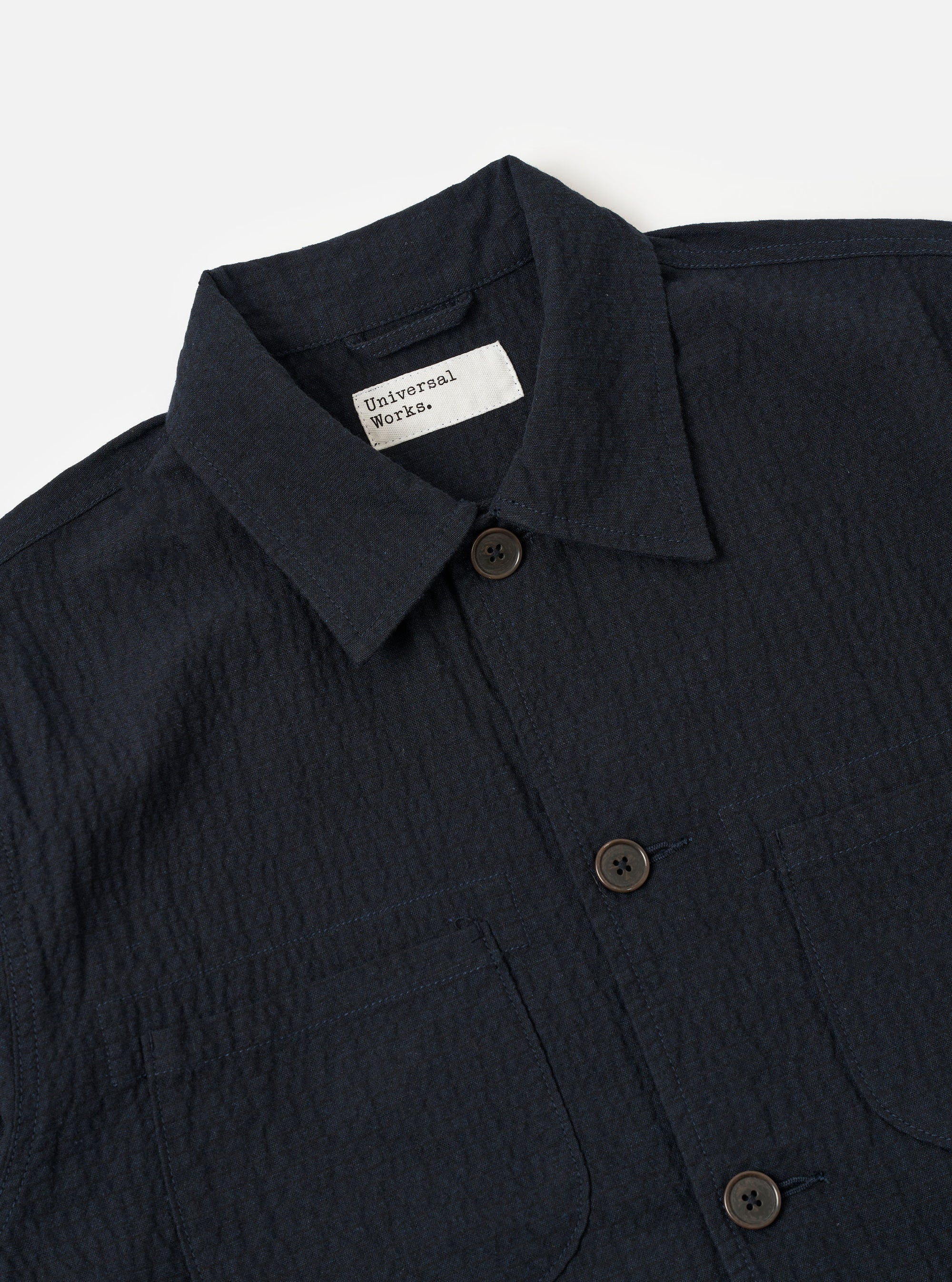 The Dark Navy Ospina Cotton Labour Jacket is shown from the back as a flat lay against a plain white background. The image highlights the jacket's structured back panel, clean seams, and a straight hem. The dark navy fabric features a visible puckered seersucker texture.