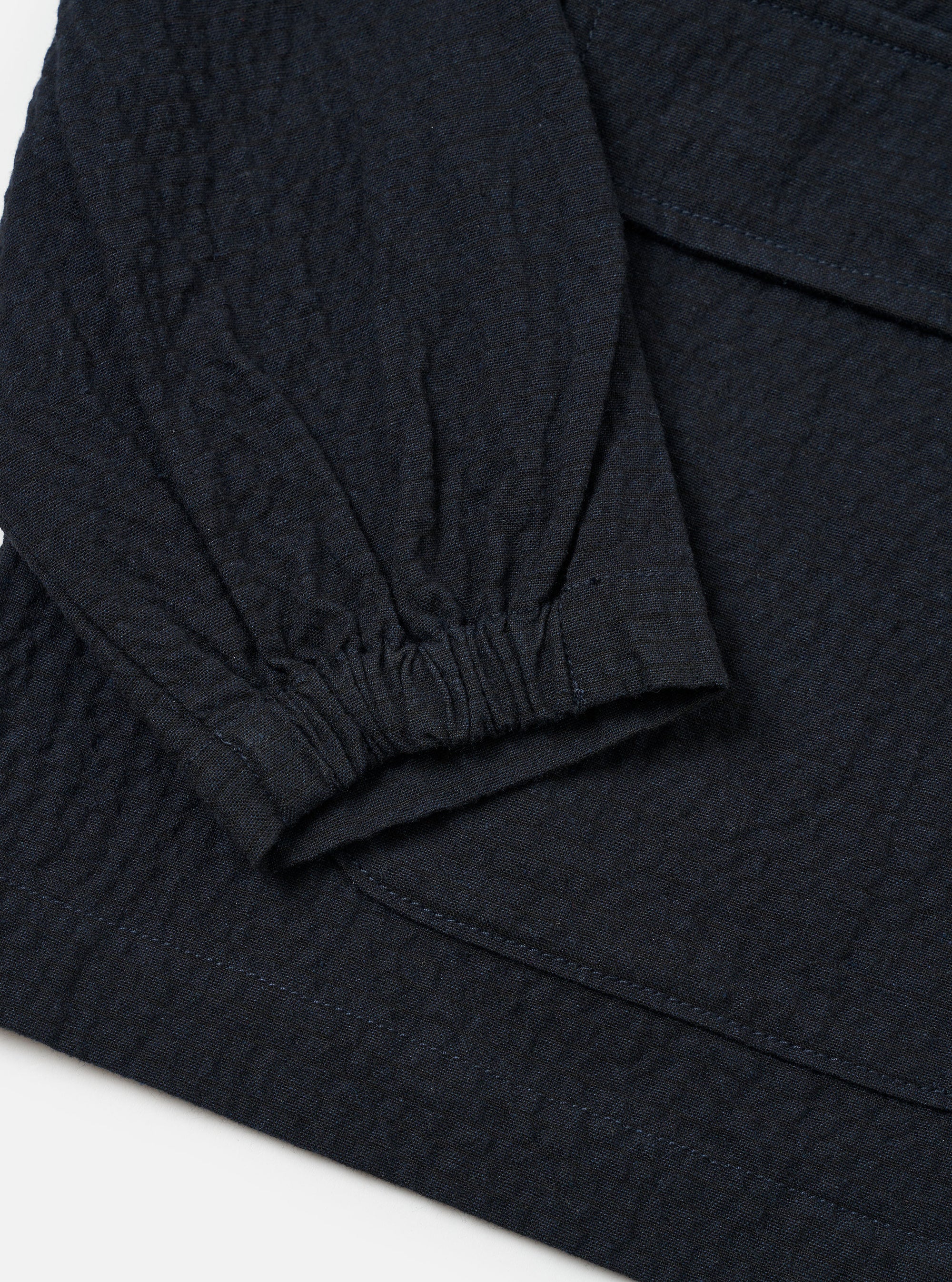 The Dark Navy Ospina Cotton Labour Jacket is shown in a detail view as a flat lay against a plain white background. The image highlights the textured seersucker finish of the navy blue fabric and the construction of the two lower flap patch pockets.