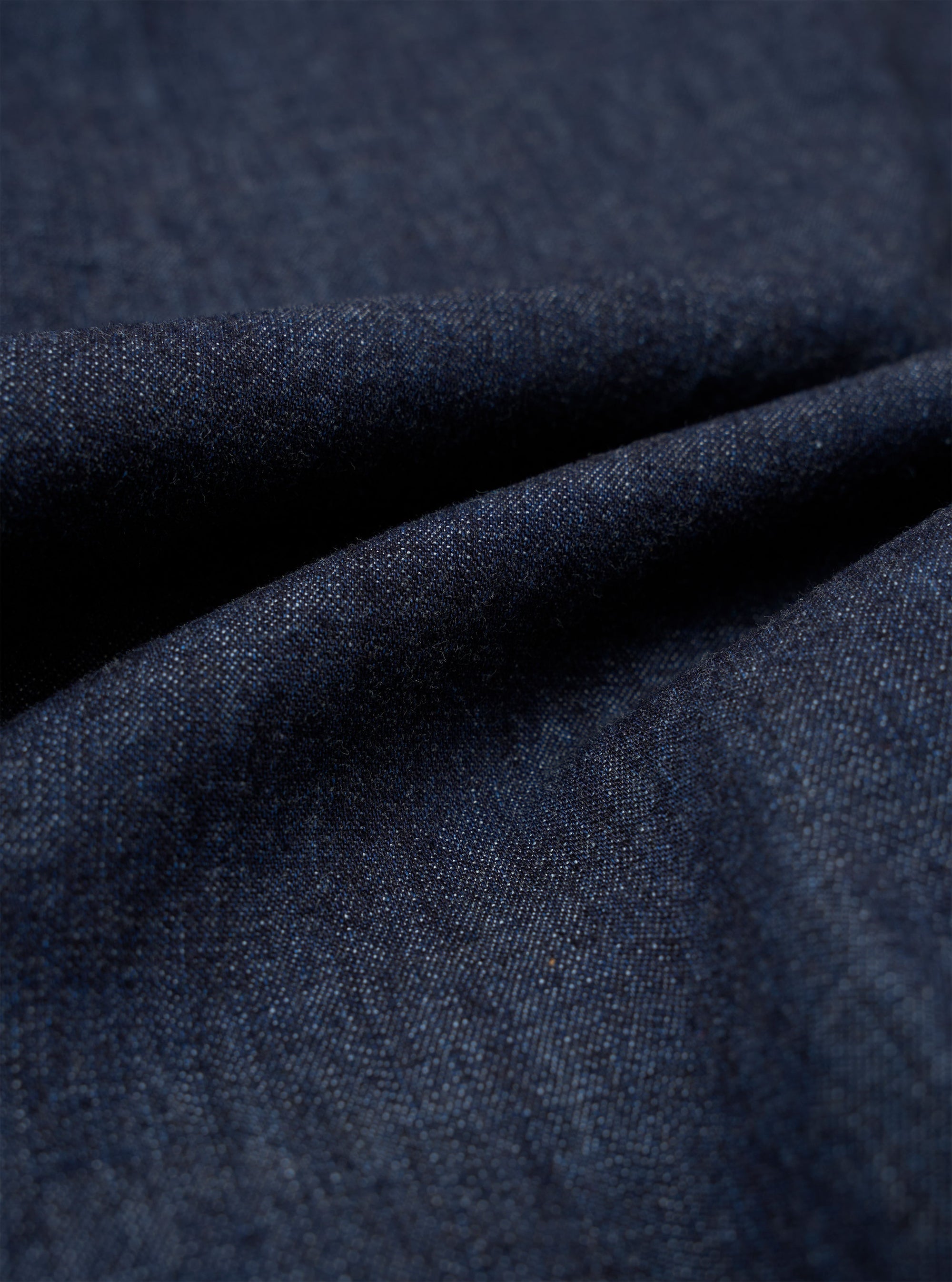 A close-up swatch of the Indigo Kaihara LT WT Denim Jungle Jacket highlights the deep indigo color and the fine diagonal weave of the lightweight Japanese denim fabric. The material fills the frame, providing a detailed view of the textile's texture and natural dye finish.