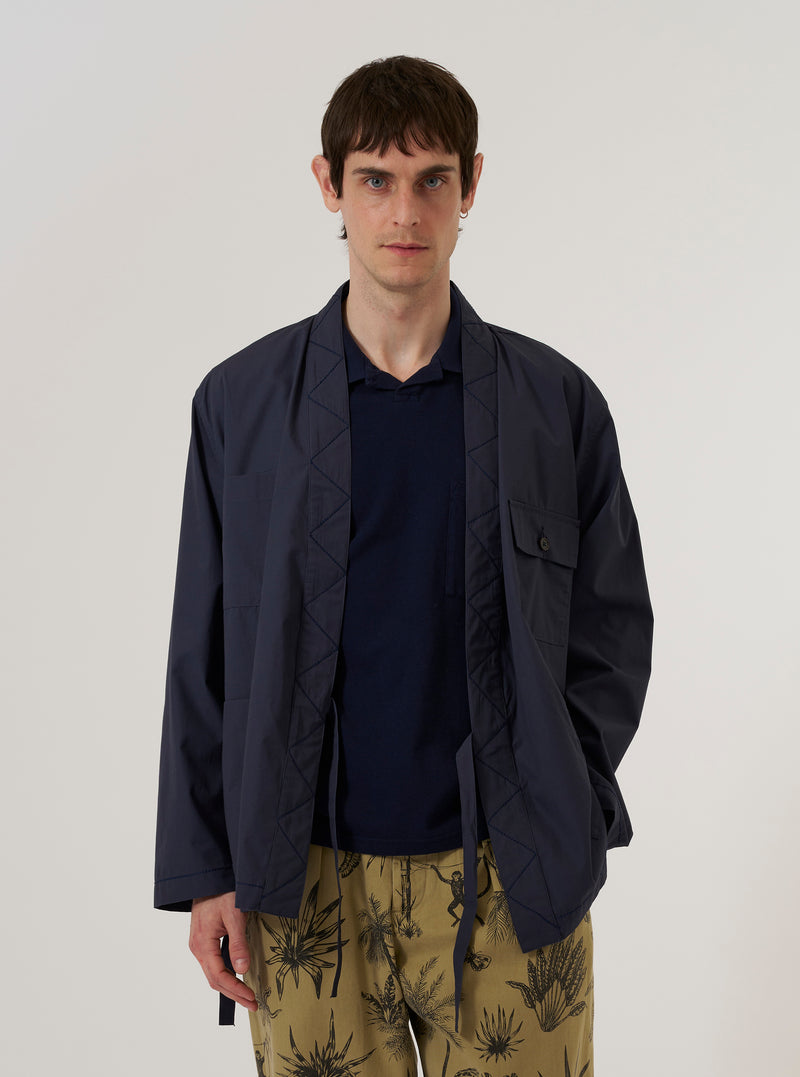A model wears the Navy Peached Poplin Osaka Work Jacket over a light-colored shirt, standing against a plain white background. The jacket features a wrap-front kimono-style design with an internal tie closure, a simple V-neckline, and two large patch pockets at the waist. The navy blue cotton poplin fabric has a smooth, matte finish.