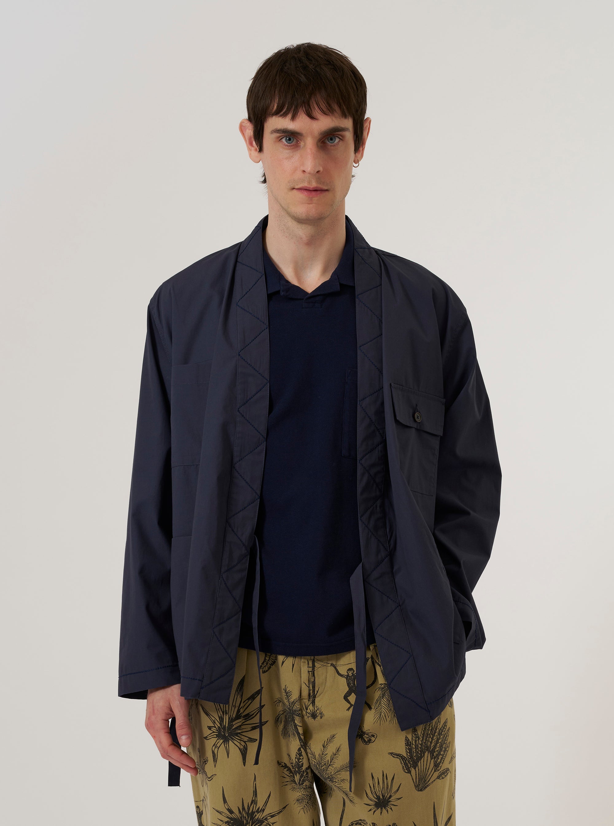 A model wears the Navy Peached Poplin Osaka Work Jacket over a light-colored shirt, standing against a plain white background. The jacket features a wrap-front kimono-style design with an internal tie closure, a simple V-neckline, and two large patch pockets at the waist. The navy blue cotton poplin fabric has a smooth, matte finish.