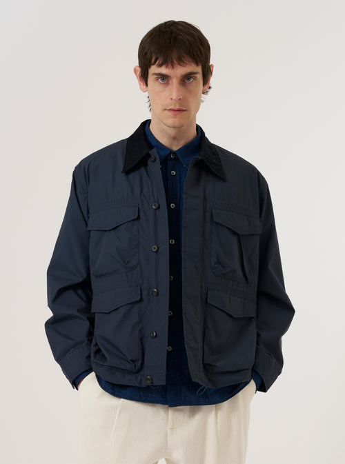 A model wears the Navy Recycled Polytech Parachute Field Jacket over a light-colored top, standing against a plain, neutral background. The jacket features a regular fit, a six-button front closure, a contrasting cord collar with a button neck strap, and four large 3D patch pockets with button flaps on the front. The fabric has a clean, utilitarian appearance in a solid navy blue.