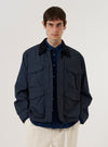A model wears the Navy Recycled Polytech Parachute Field Jacket over a light-colored top, standing against a plain, neutral background. The jacket features a regular fit, a six-button front closure, a contrasting cord collar with a button neck strap, and four large 3D patch pockets with button flaps on the front. The fabric has a clean, utilitarian appearance in a solid navy blue.