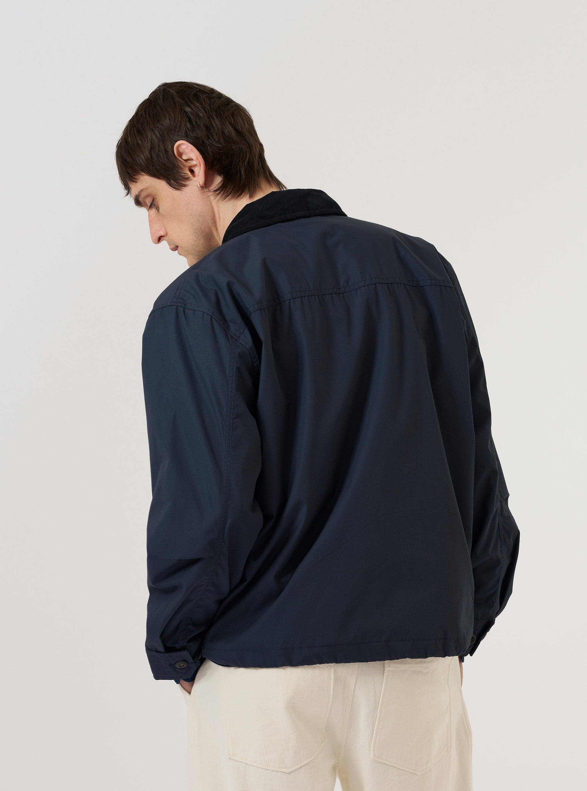 A model wears the Navy Recycled Polytech Parachute Field Jacket over a light-colored long-sleeve shirt, shown from a rear three-quarter angle against a plain white background. The jacket features a regular fit, a straight hem, and a contrasting cord collar. The navy blue fabric has a smooth, technical finish. No mannequin is present in the image.