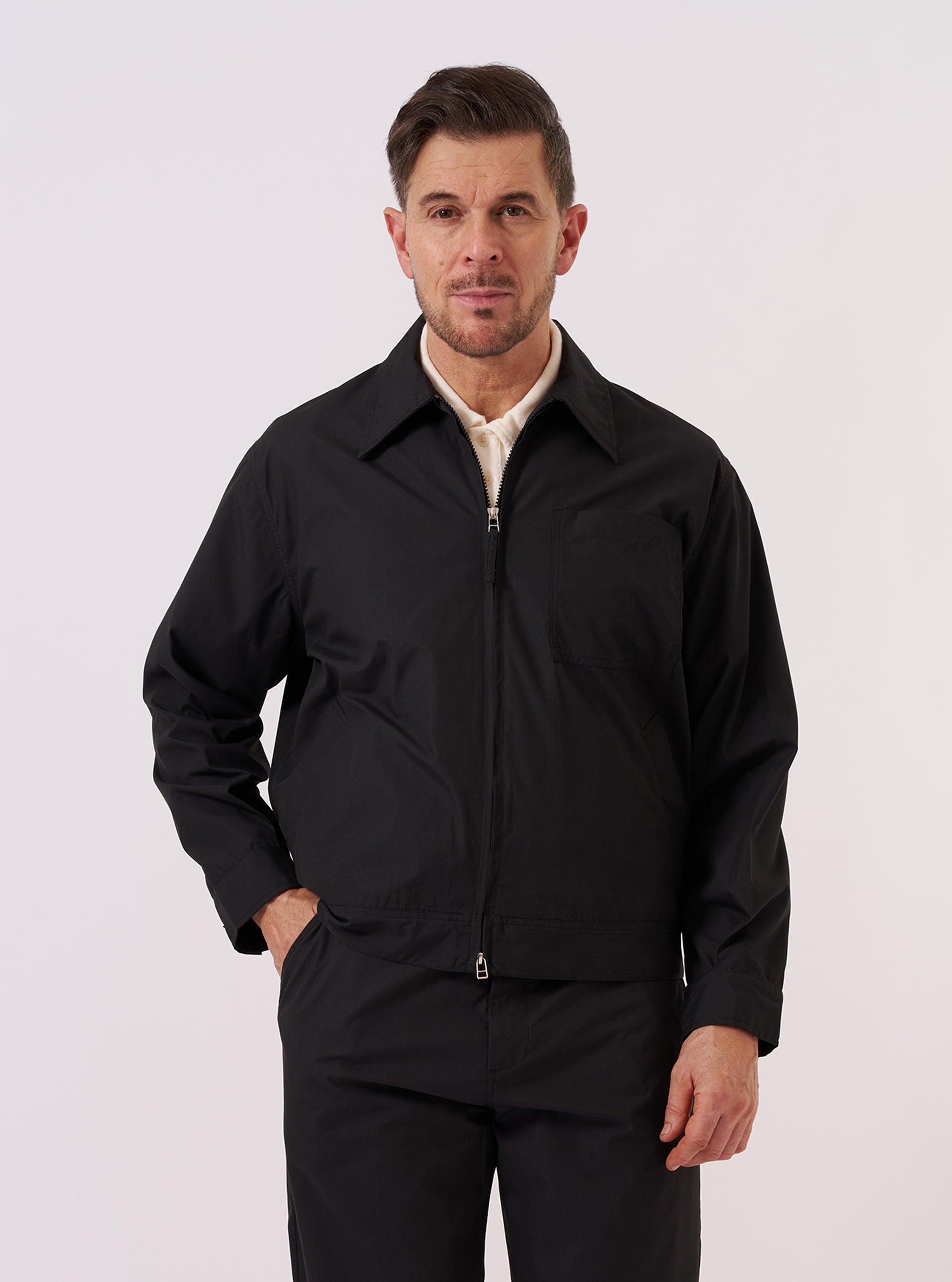 A model wears the Black Recycled Polytech Rose Bowl Jacket against a plain, neutral background. The jacket features a boxy fit, a central zip front, and a slightly oversized collar. Prominent details include a single large patch pocket on the chest and two welt pockets at the waist. The black recycled polytech fabric has a smooth, technical appearance.