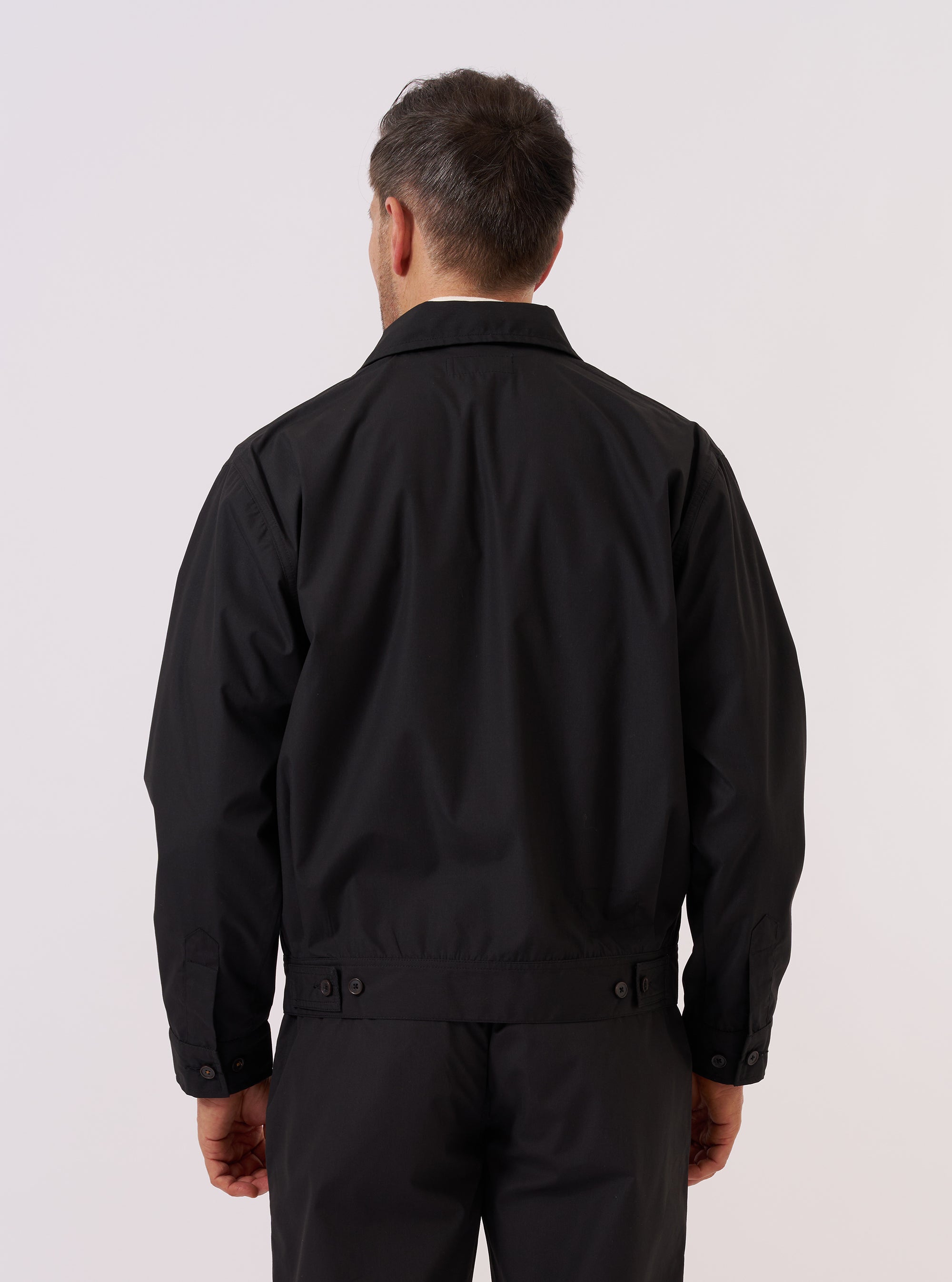 A model is wearing the Black Recycled Polytech Rose Bowl Jacket, shown from a rear three-quarter angle against a plain white background. The jacket features a boxy fit, an oversized collar, and pleats on the back and cuffs. The black fabric has a smooth, utilitarian appearance. No mannequin is present in the image.