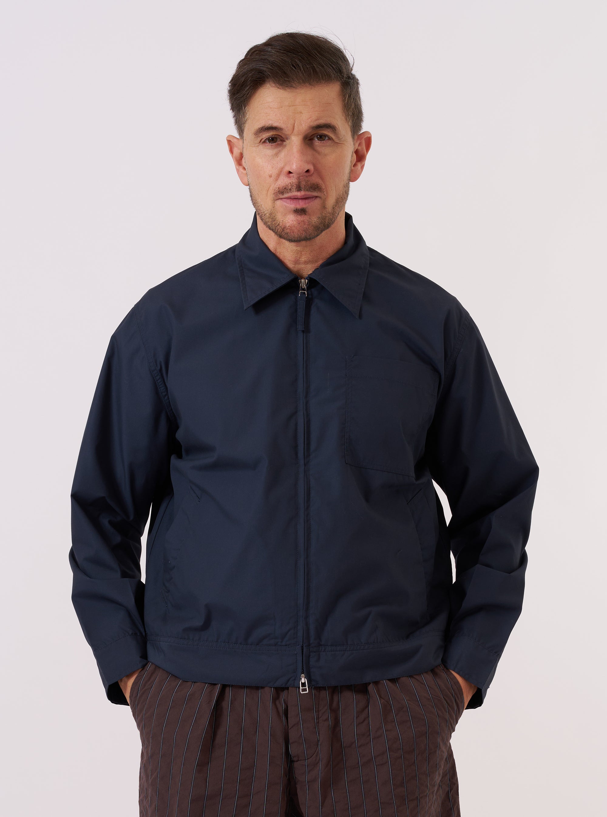 A model wears the Navy Recycled Polytech Rose Bowl Jacket over a light-colored shirt, standing against a plain, neutral studio background. The jacket features a short, boxy fit with a central two-way zip closure and a slightly oversized collar. Prominent details include a large patch pocket on the left chest and two slanted welt pockets at the waist. The smooth, dark navy fabric has a clean, utilitarian appearance. No mannequin is present in the image.