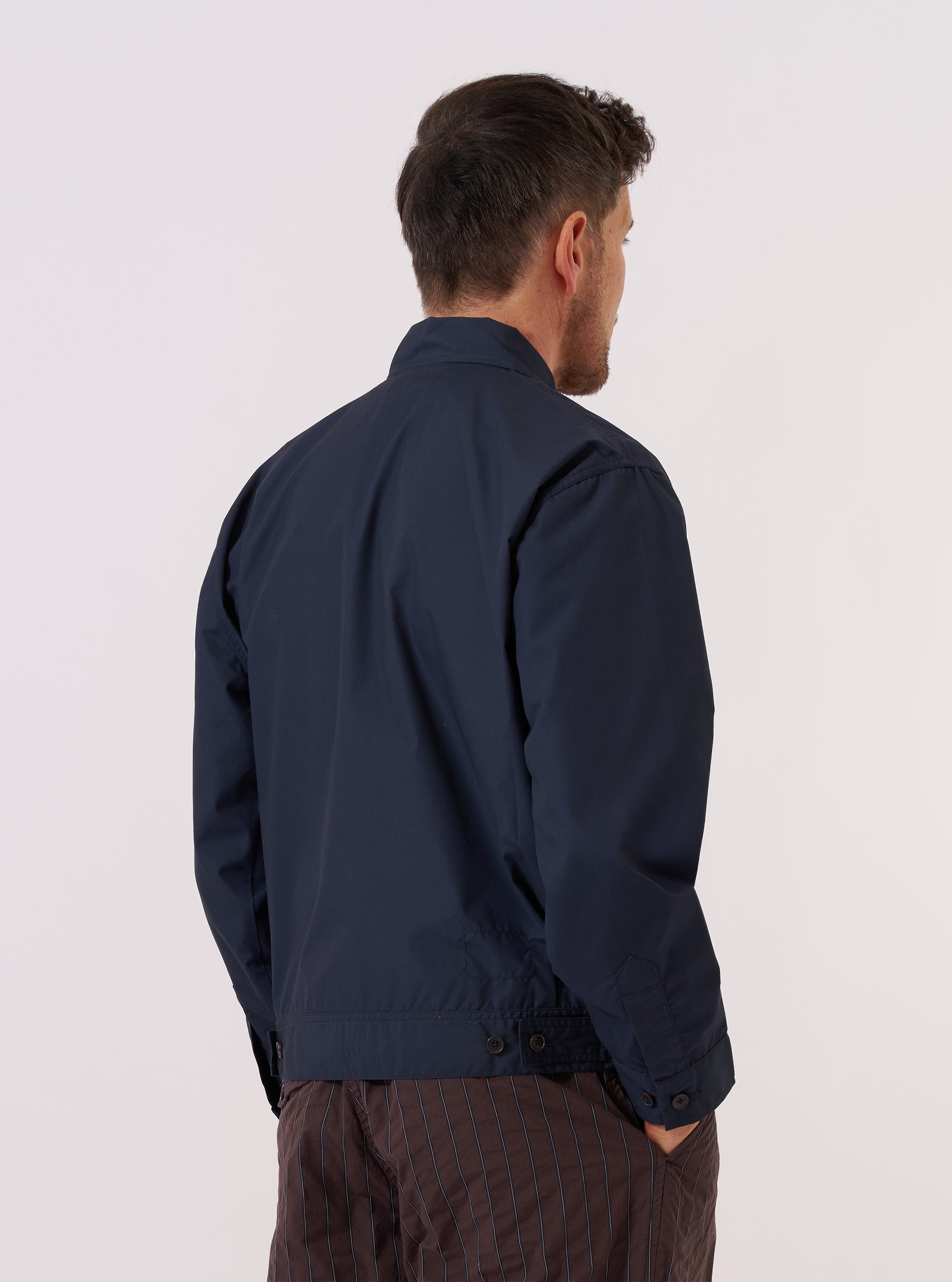 A model wears the Navy Recycled Polytech Rose Bowl Jacket over a light-colored shirt, shown from a side-angle view against a plain white background. The jacket features a classic turn-down collar, a central button-down closure, and a large patch pocket on the front. The navy fabric has a smooth, technical finish.