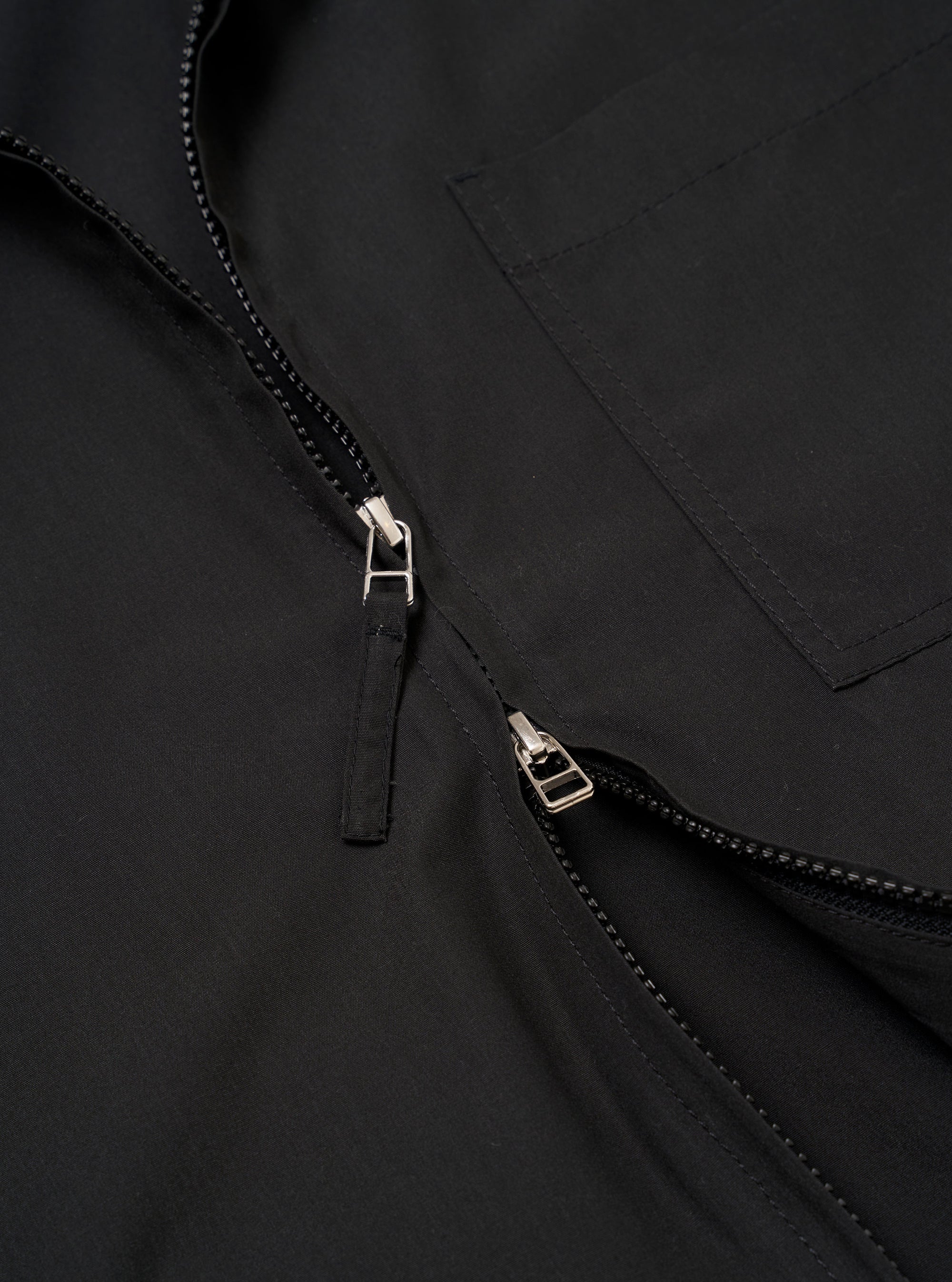 The Black Recycled Polytech Rose Bowl Jacket is shown in a close-up detail shot as a flat lay against a plain white background. The image highlights the technical texture of the black fabric and the construction of the front pockets and the central two-way zipper closure.