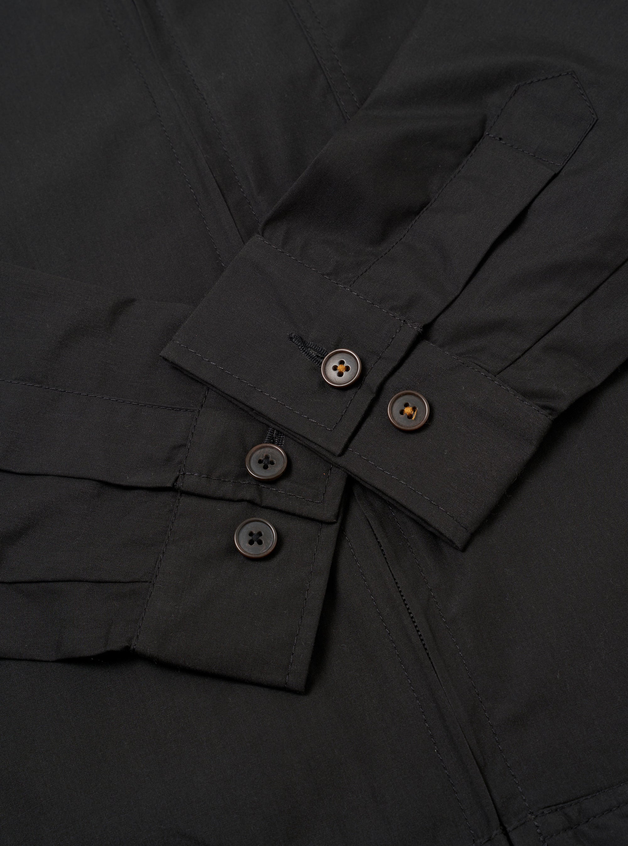 The Black Recycled Polytech Rose Bowl Jacket is shown from the back as a flat lay against a plain white background. The image highlights the jacket's simple construction, featuring a straight hem with adjustable button tabs on either side and vertical pleats extending from the shoulder yoke to the hem.