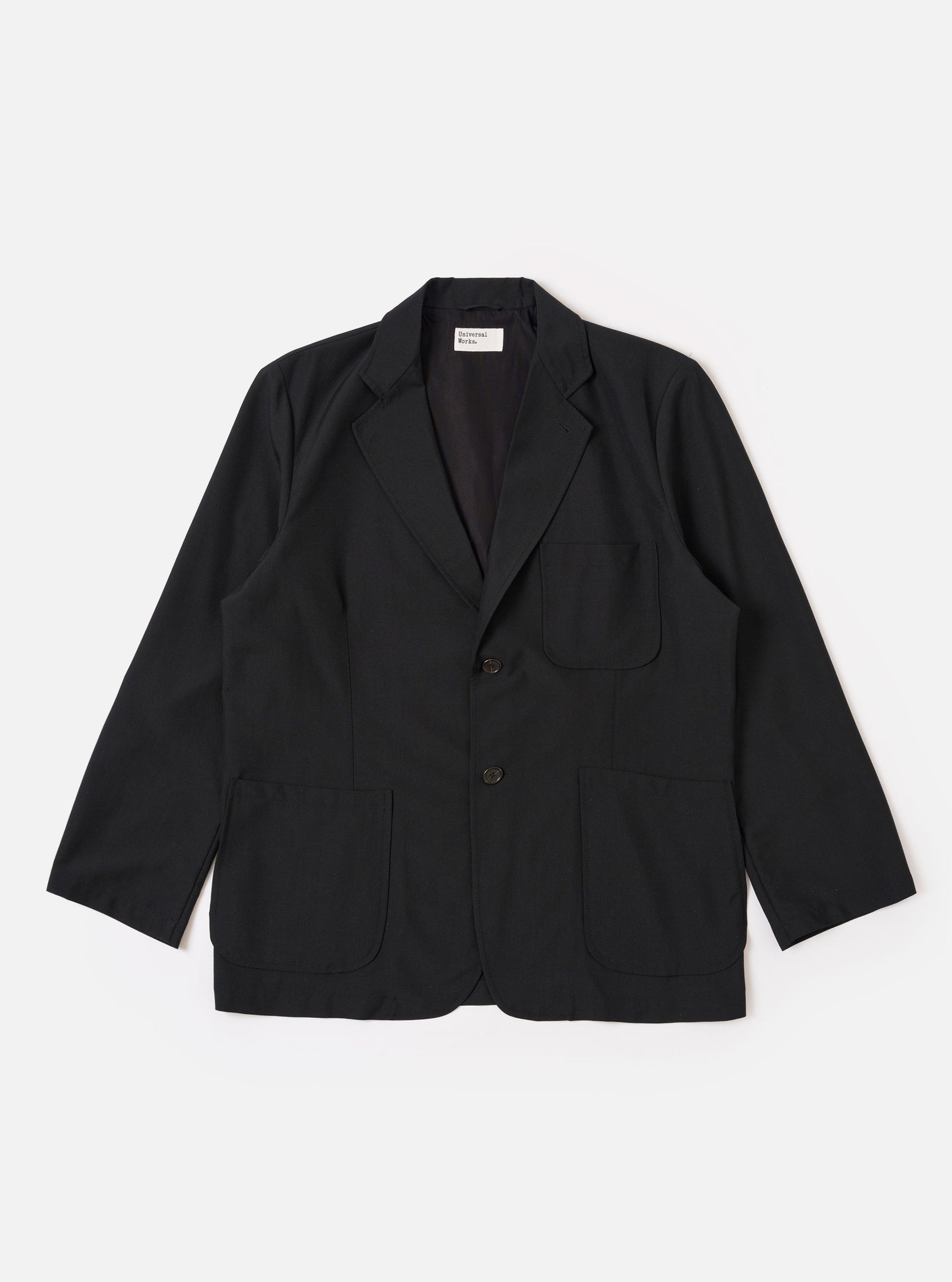 The Black Tropical Suiting Two Button Jacket is shown as a flat lay against a white background. The jacket features a relaxed fit with a long, wide notched lapel, two large lower patch pockets, and a single chest patch pocket. The two-button closure is visible at the center, and the fabric has a smooth, lightweight appearance.