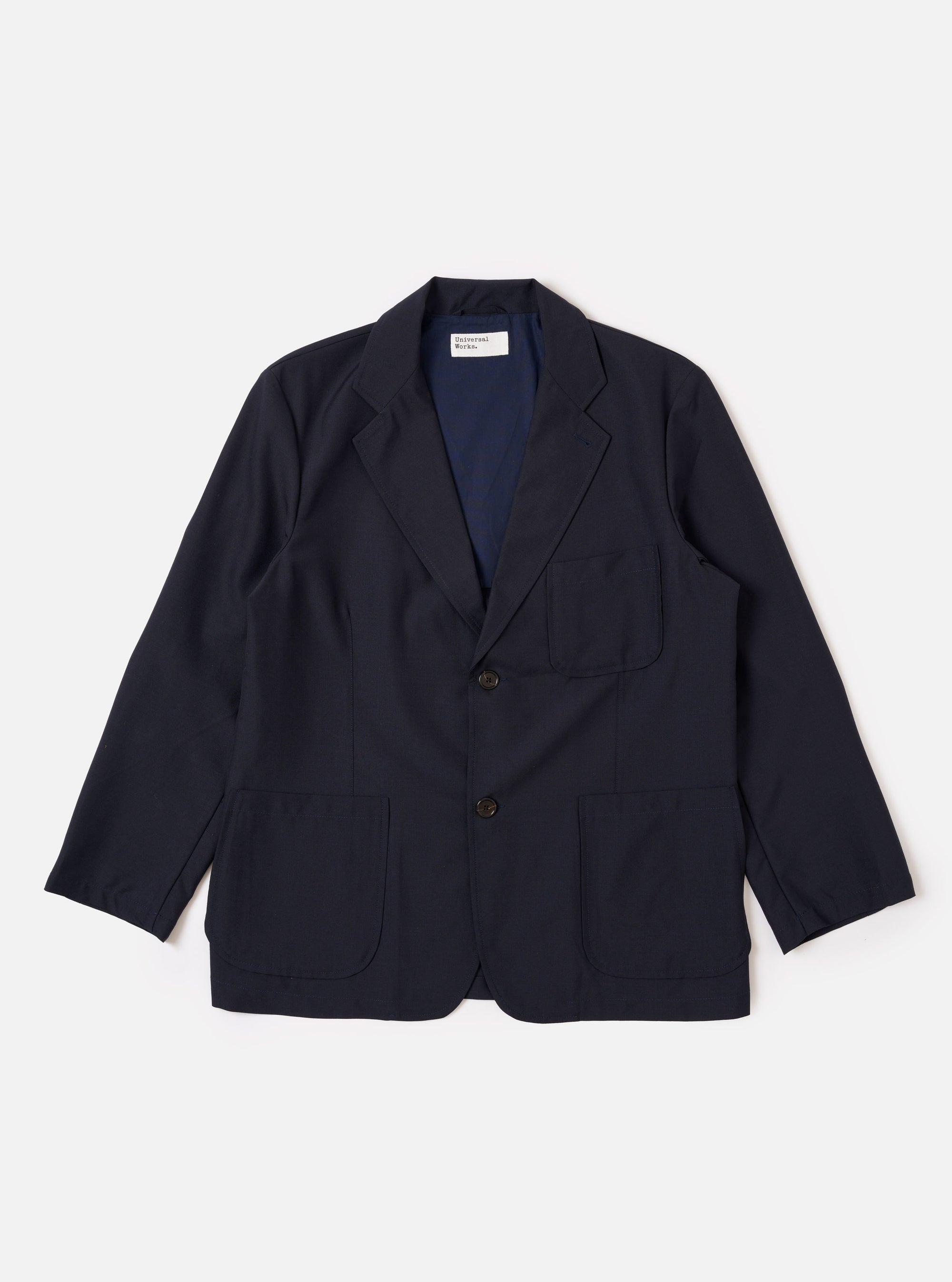 The Navy Tropical Suiting Two Button Jacket is shown as a flat lay against a white background. The jacket features a two-button front closure, a notched lapel with pick-stitching, and three front pockets, including two large patch pockets at the waist and one at the chest. The fabric is a smooth, lightweight navy tropical suiting material.