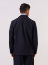 A model wears the Navy Tropical Suiting Two Button Jacket, shown from the back against a plain white background. The jacket features structured shoulders, a clean tailored fit with vertical seams, and a central rear vent at the hem. The garment is made from a smooth navy blue tropical suiting fabric and is worn over a light-colored top.