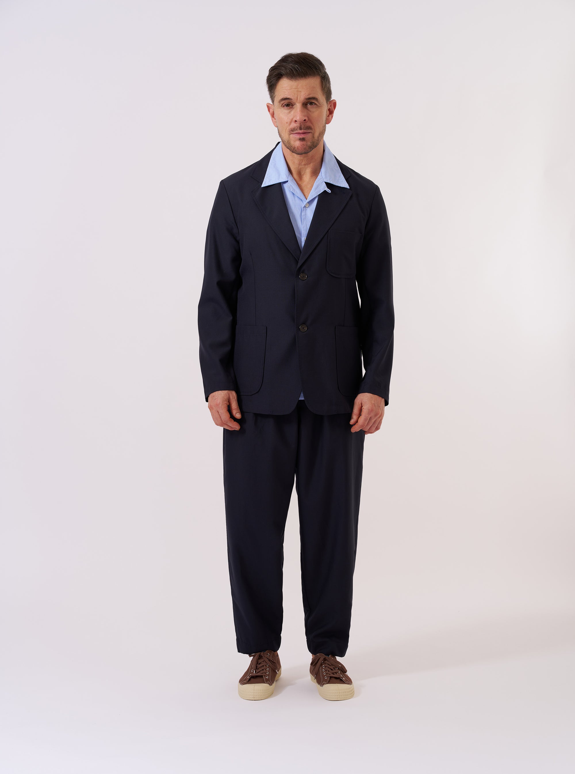 The Navy Tropical Suiting Two Button Jacket is displayed as a flat lay against a plain white background. The jacket features a relaxed fit with a two-button front closure, a wide notch lapel, and three front patch pockets, including one at the chest and two at the waist. The navy blue fabric is a lightweight tropical wool blend with a smooth, refined texture.
