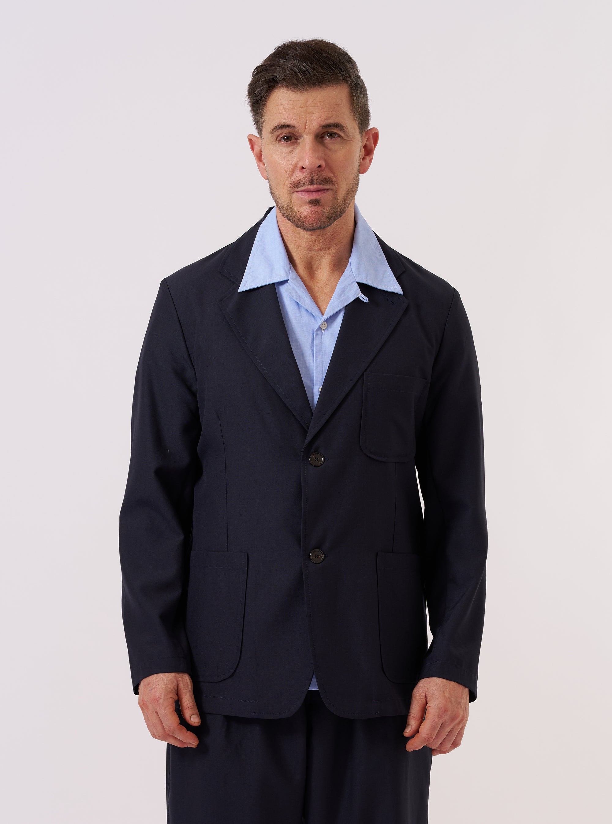 A model wears the Navy Tropical Suiting Two Button Jacket over a light-colored shirt, standing against a plain, neutral studio background. The jacket features a two-button front closure, classic notch lapels, and two large patch pockets at the waist. The navy wool-blend fabric has a smooth, tailored finish.